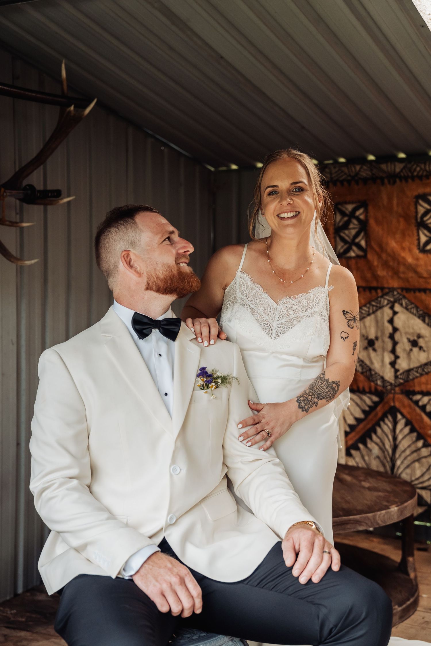 Smiling bride and groom in white tuxedo sharing a joyful moment in a rustic venue.
