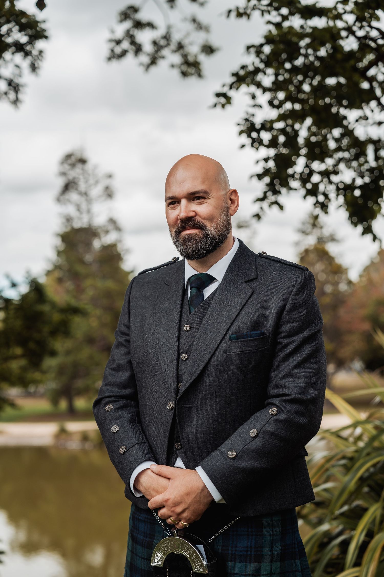 Bald groom with beard wearing traditional Scottish kilt and jacket poses by a pond.