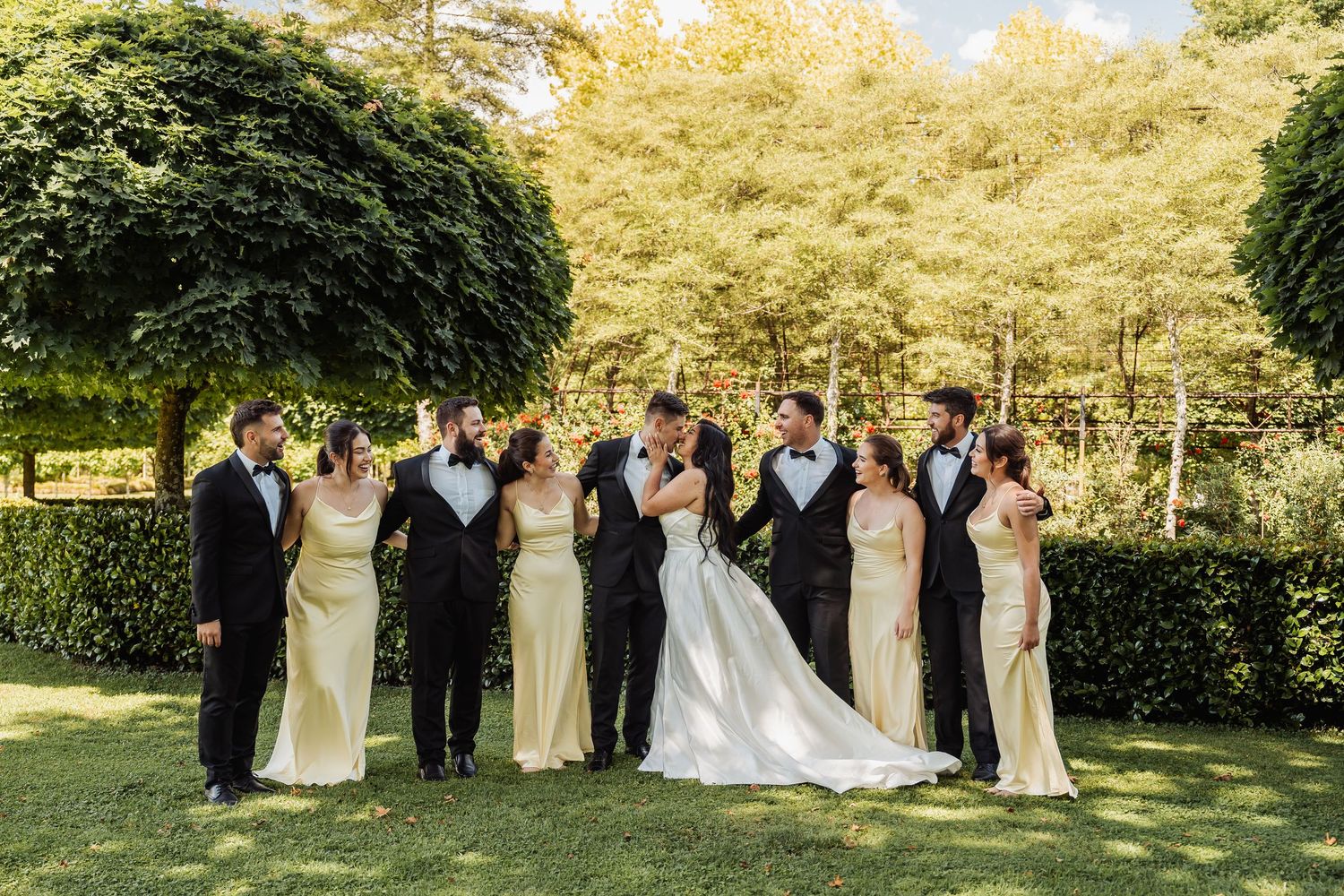 Wedding party poses outdoors, bride and groom kissing, bridesmaids in gold gowns and groomsmen in black tuxedos.