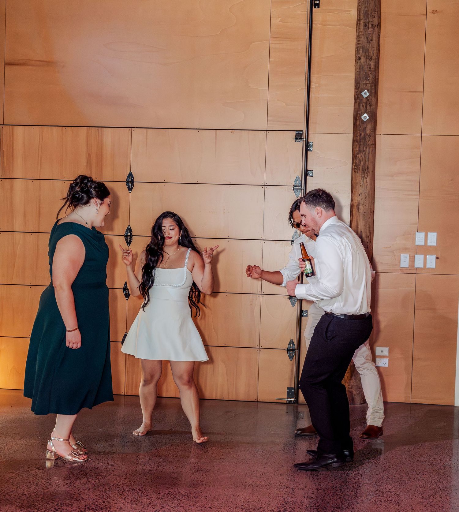 Bride in short white dress dances energetically with guests inside modern wooden-walled wedding reception venue.