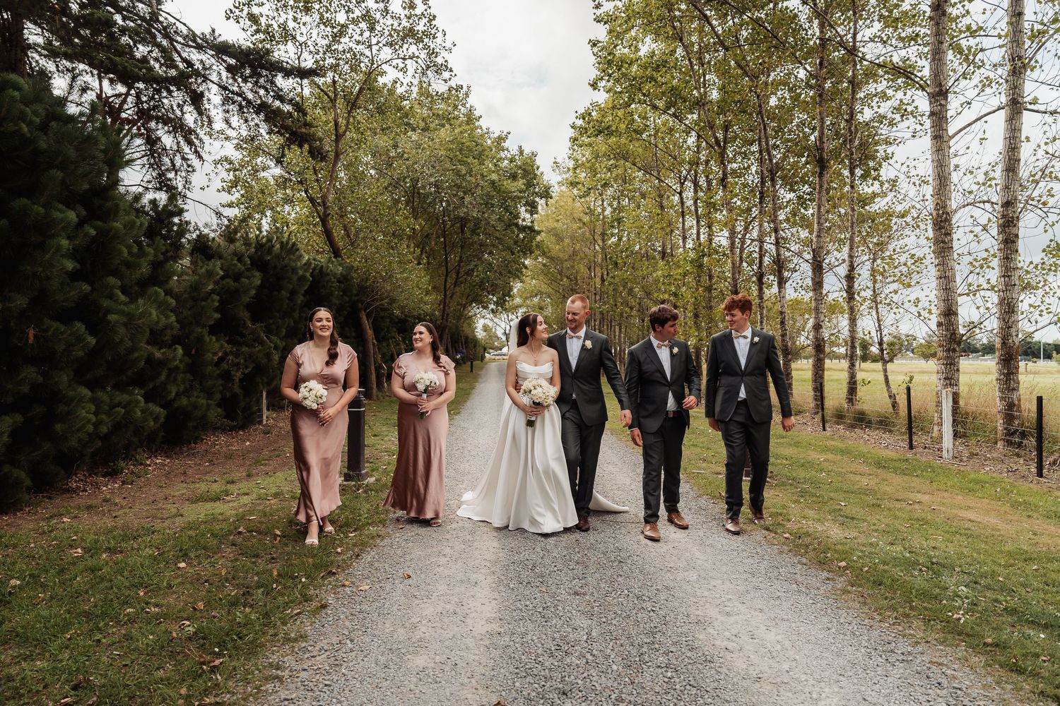 Wedding party walking together down a scenic tree-lined gravel path with bridesmaids in dusty rose gowns.