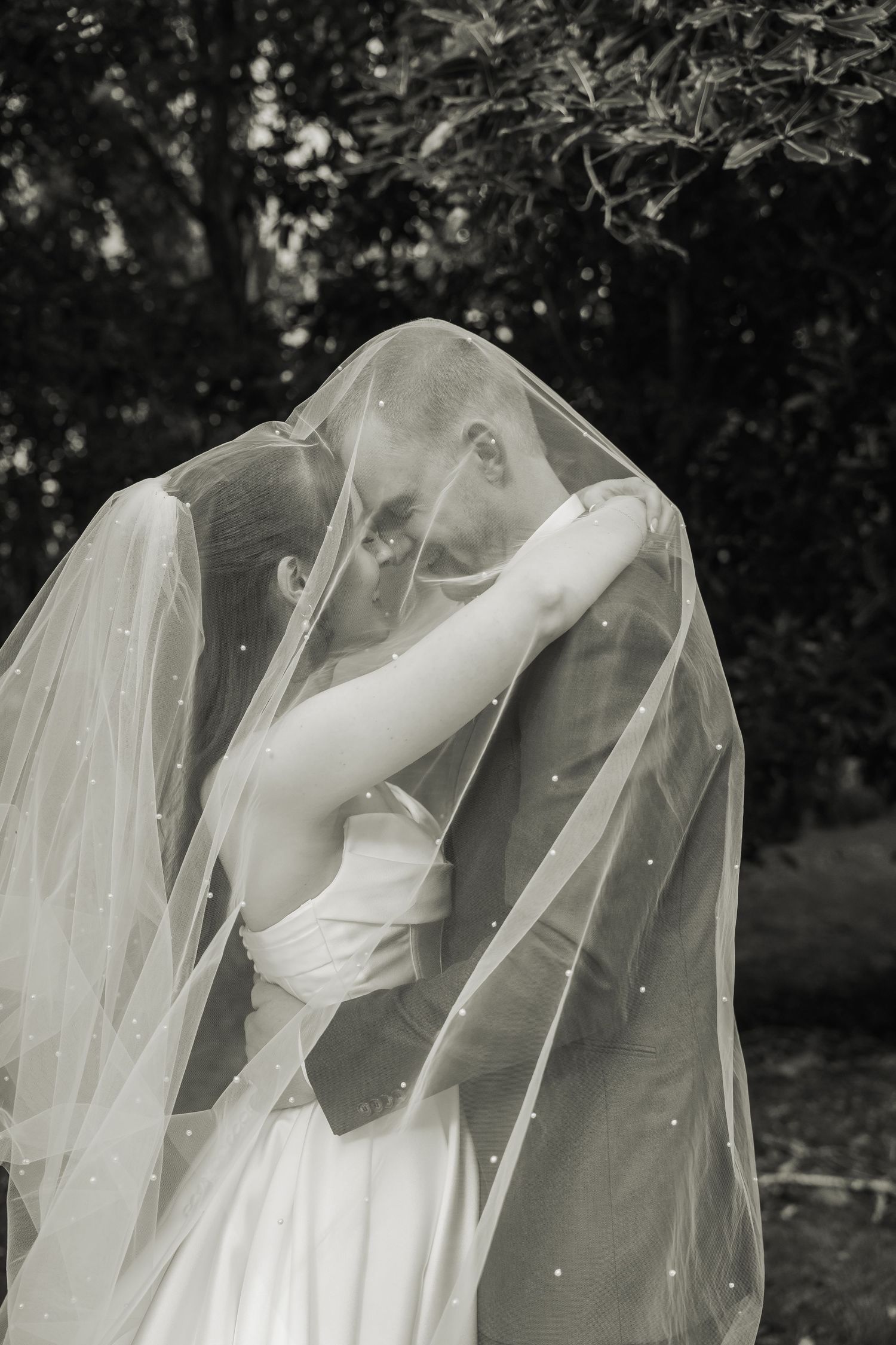 Black and white photo of bride and groom kissing beneath flowing pearl-dotted veil in garden setting.