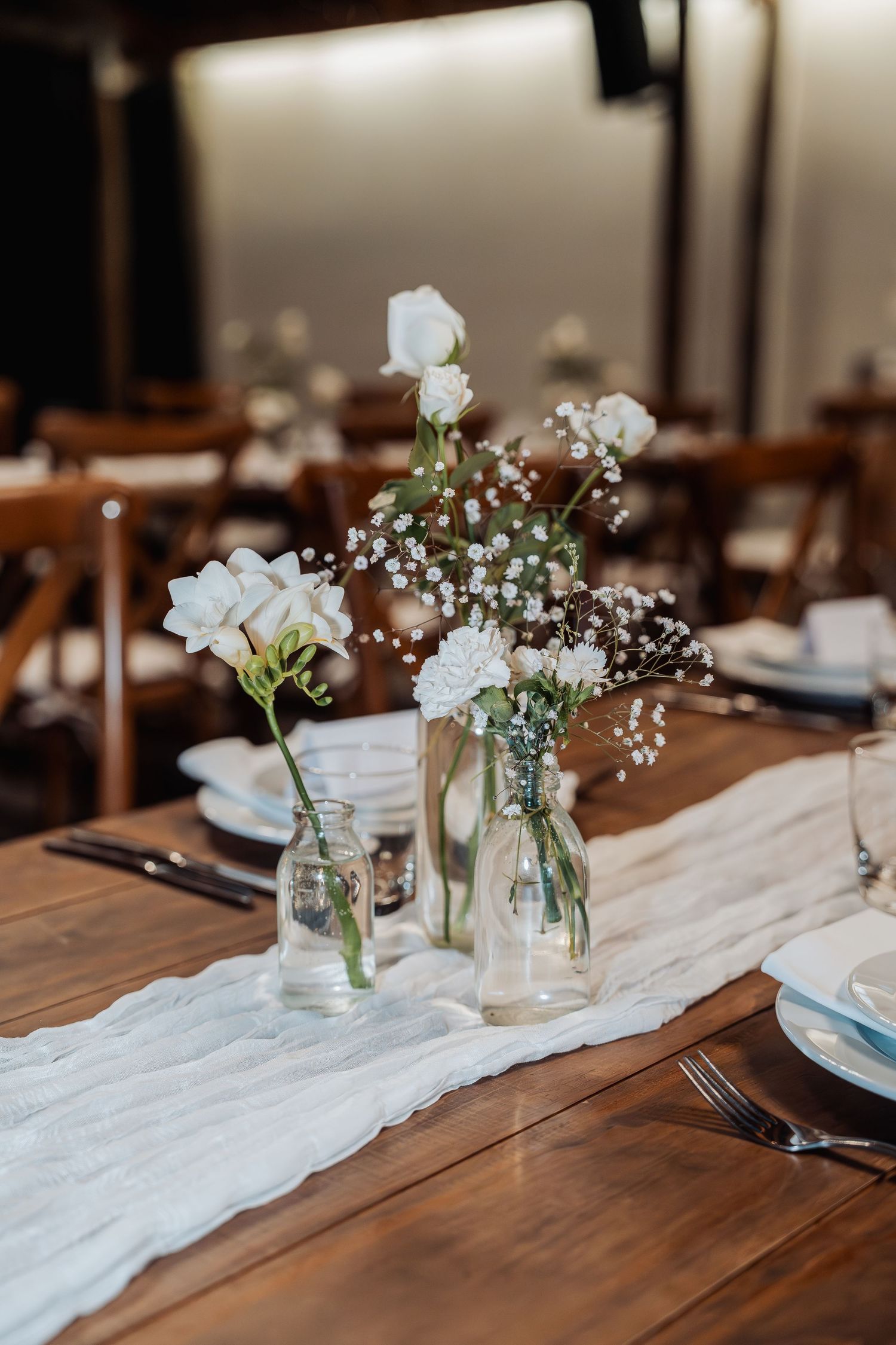Elegant wedding reception table with white floral arrangements in glass vases on a rustic wooden table with white linen runner.