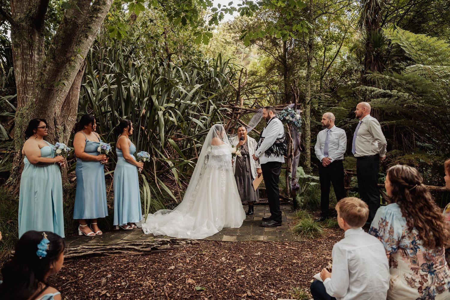 Outdoor woodland wedding ceremony with bridesmaids in blue dresses and bride in white gown with veil.