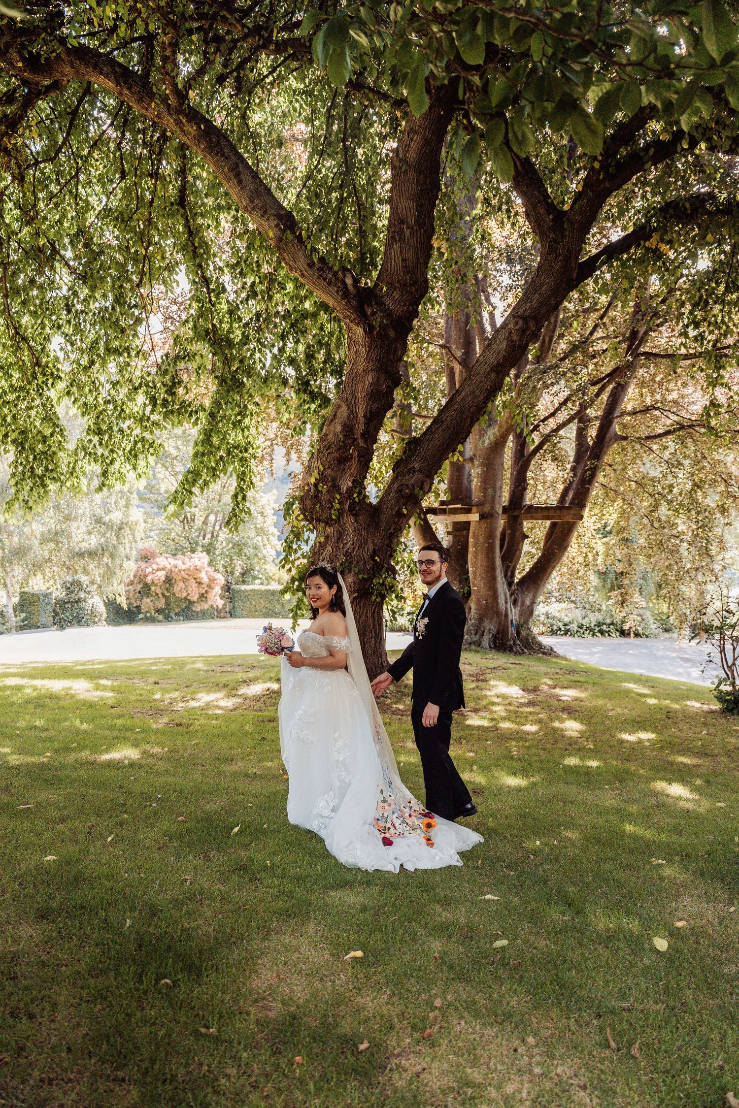 Bride and groom hold hands under large tree on green lawn at outdoor wedding venue.