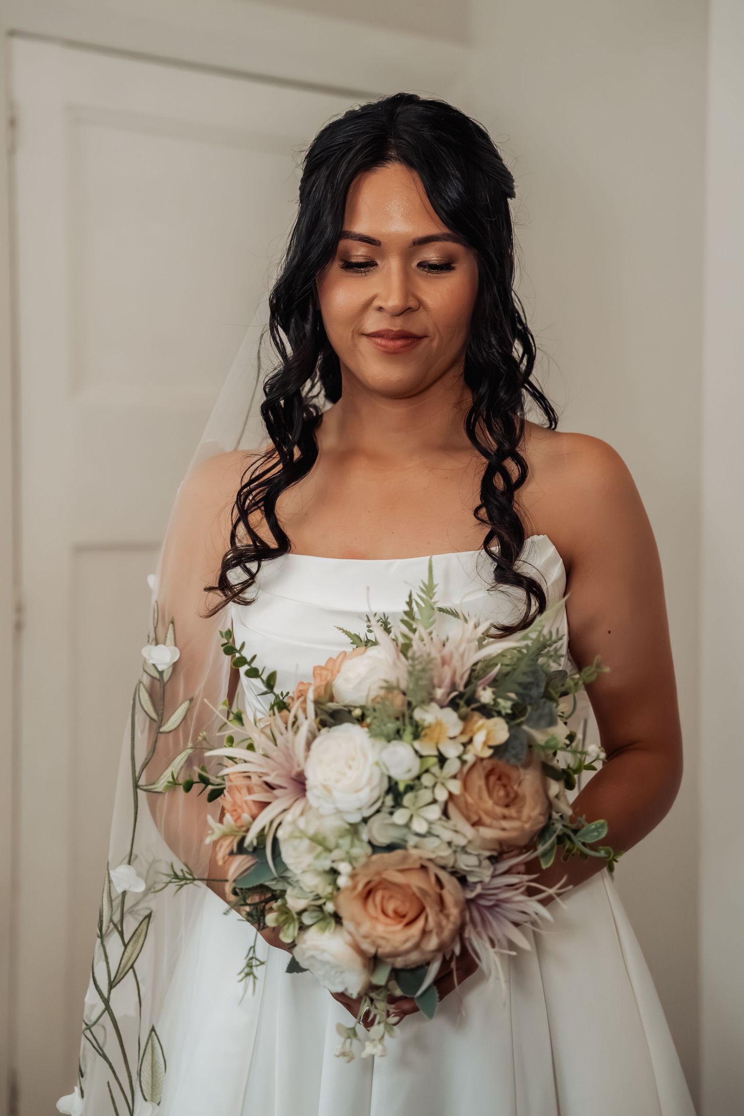 Bride in strapless white gown looks down at elegant bouquet of blush roses and white flowers with greenery.