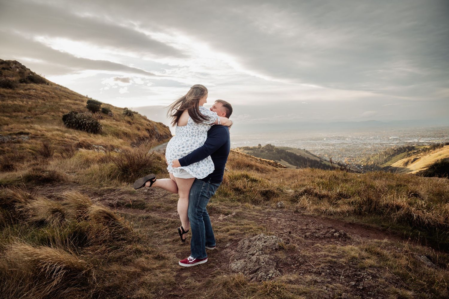 Couple sharing a romantic moment on a hilltop, man lifting woman against a dramatic cloudy sky backdrop.