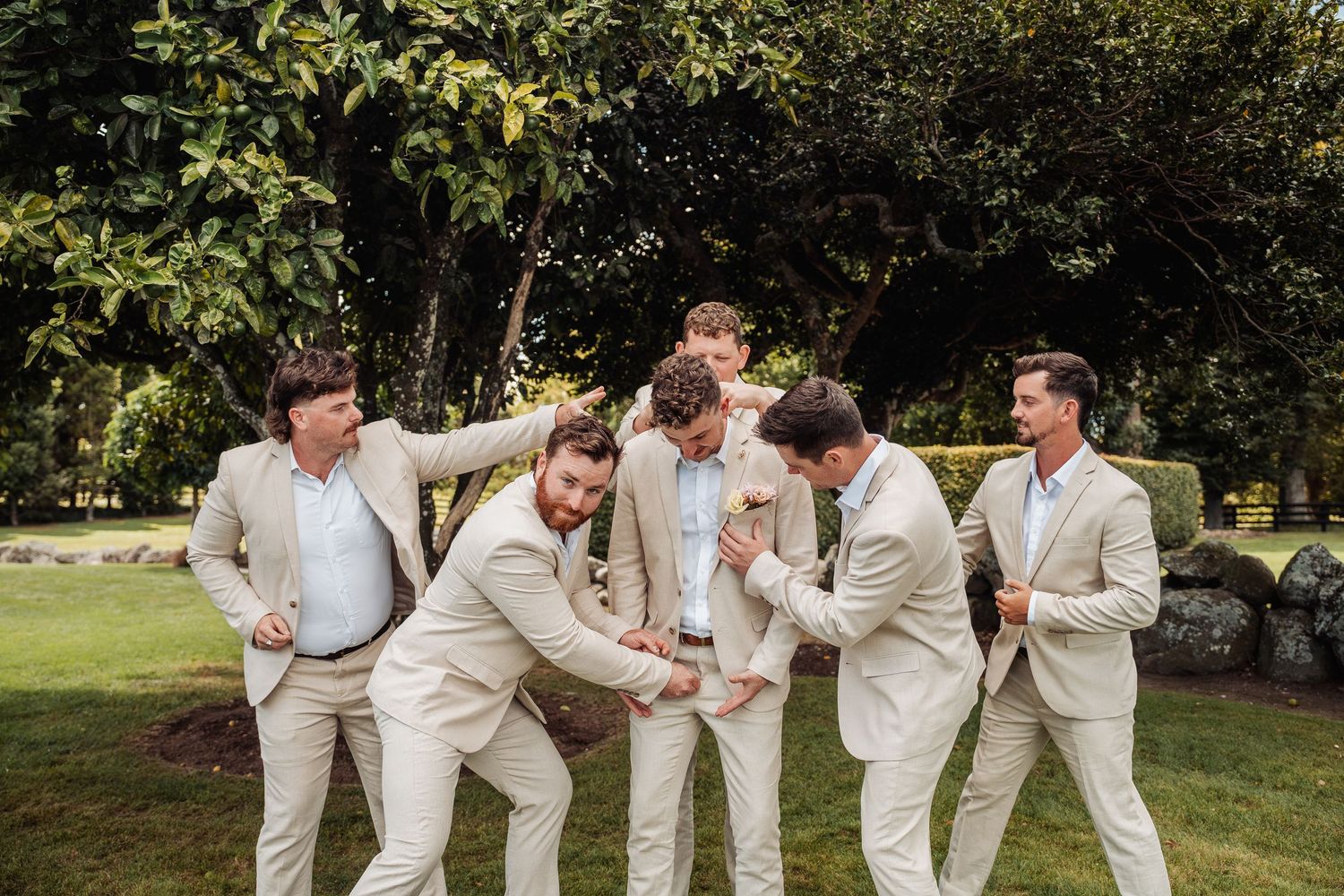 Groomsmen in beige suits laughing and playing around outdoors on a sunny day.
