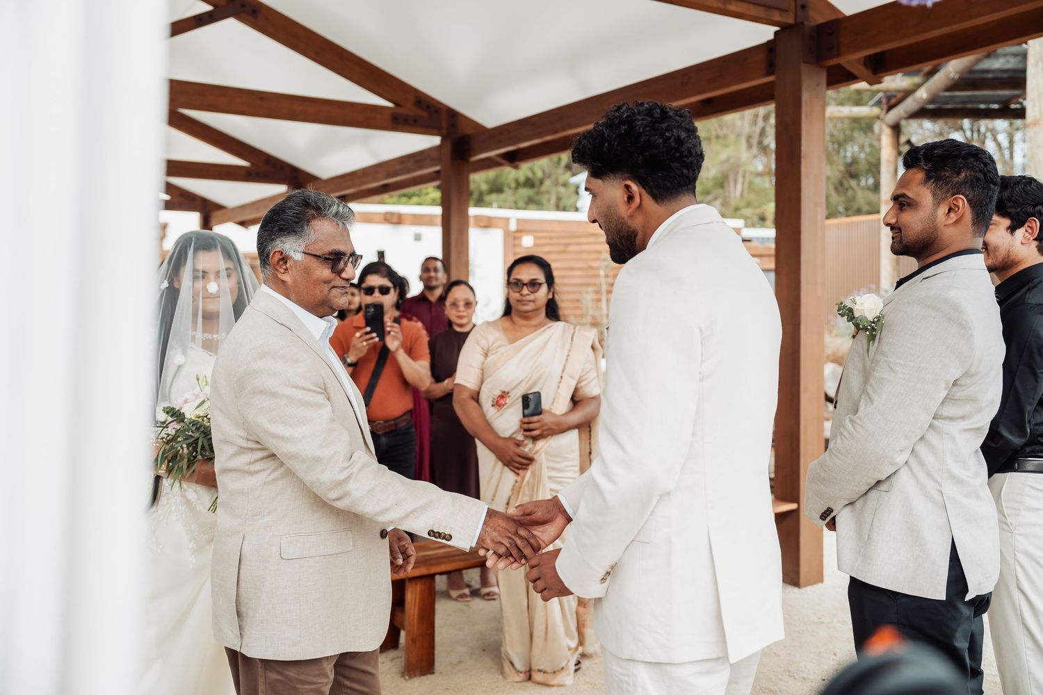 Groom shaking hands with father during outdoor wedding ceremony while guests look on.
