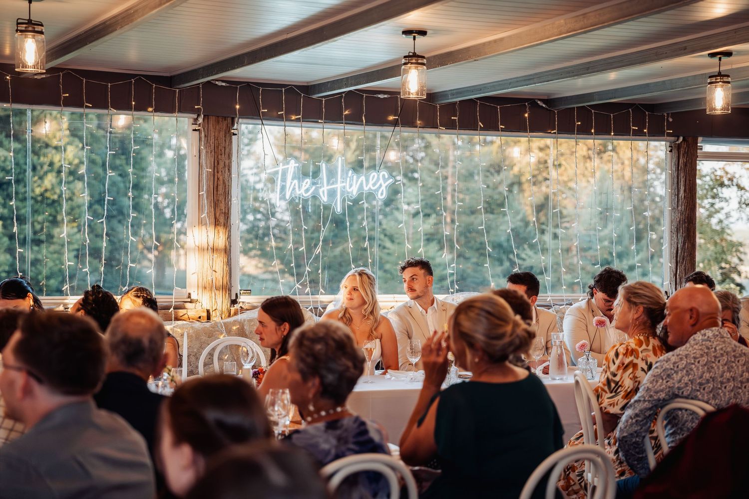 Wedding reception guests seated at tables beneath string lights with neon sign backdrop.