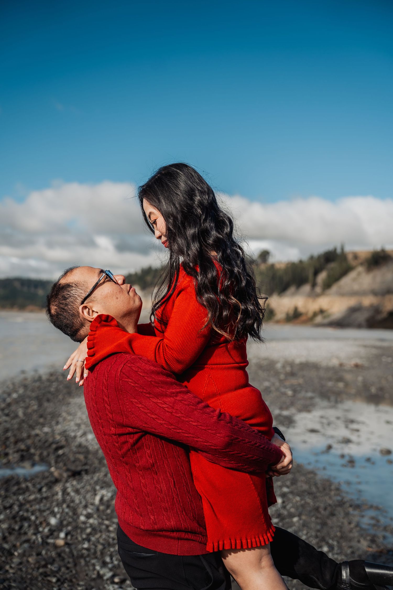 Man in red sweater lifts smiling woman in red dress on a rocky beach under a blue sky.
