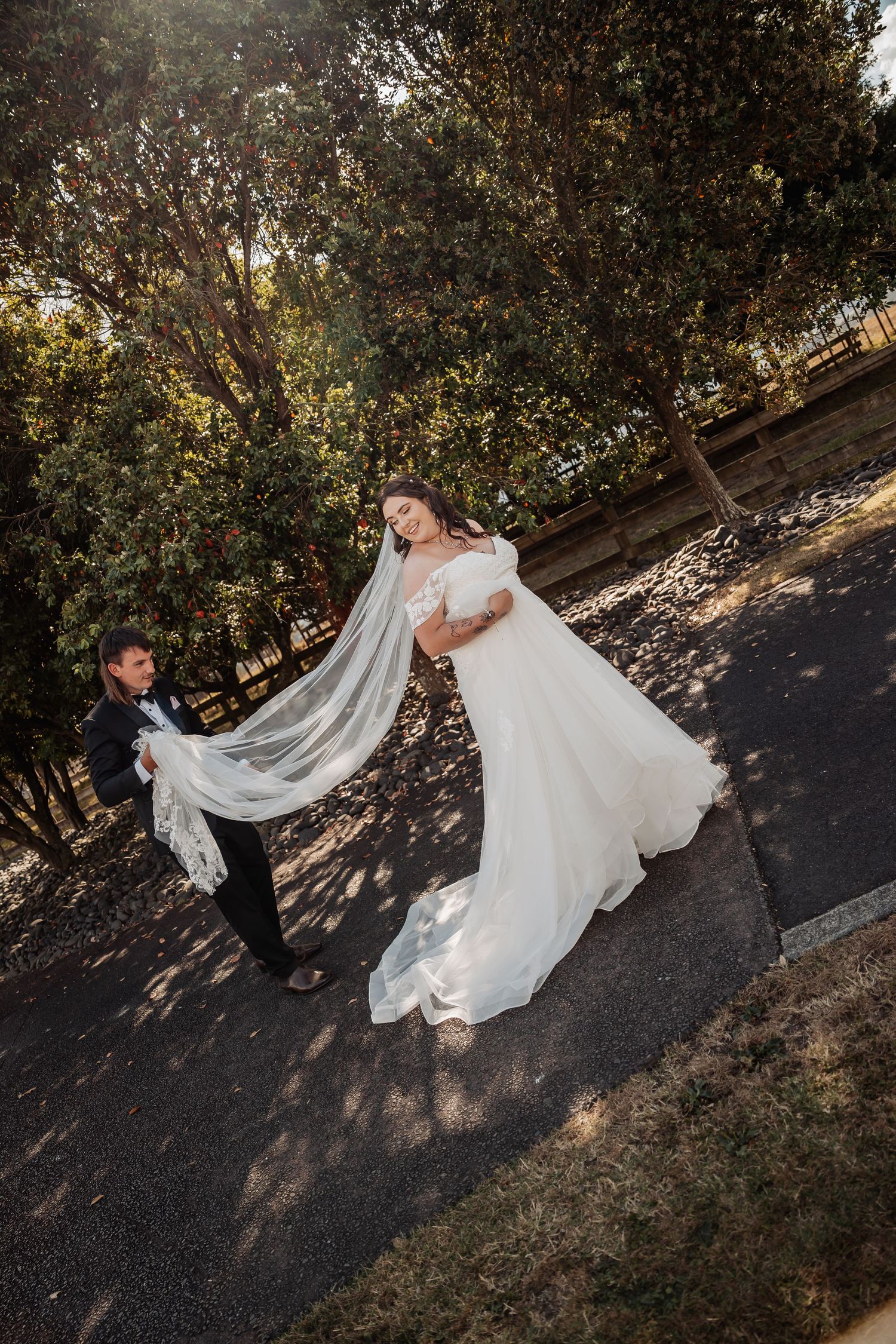 Groom holding bride's veil as she poses in white gown outdoors in a tree-lined park during golden hour.