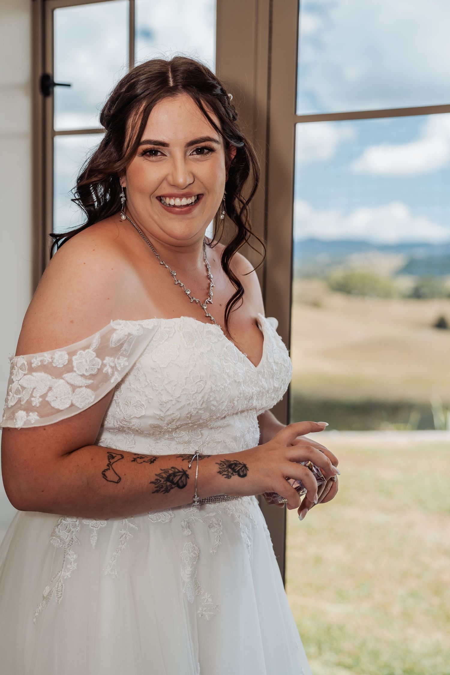 Smiling bride in off-shoulder lace wedding dress with tattoos and jewelry, posing by a window with scenic countryside views.