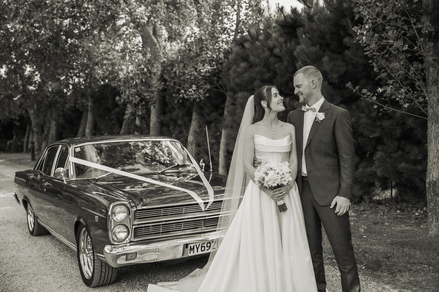 Black and white wedding photo of bride and groom posing beside a classic vintage car decorated with ribbon.