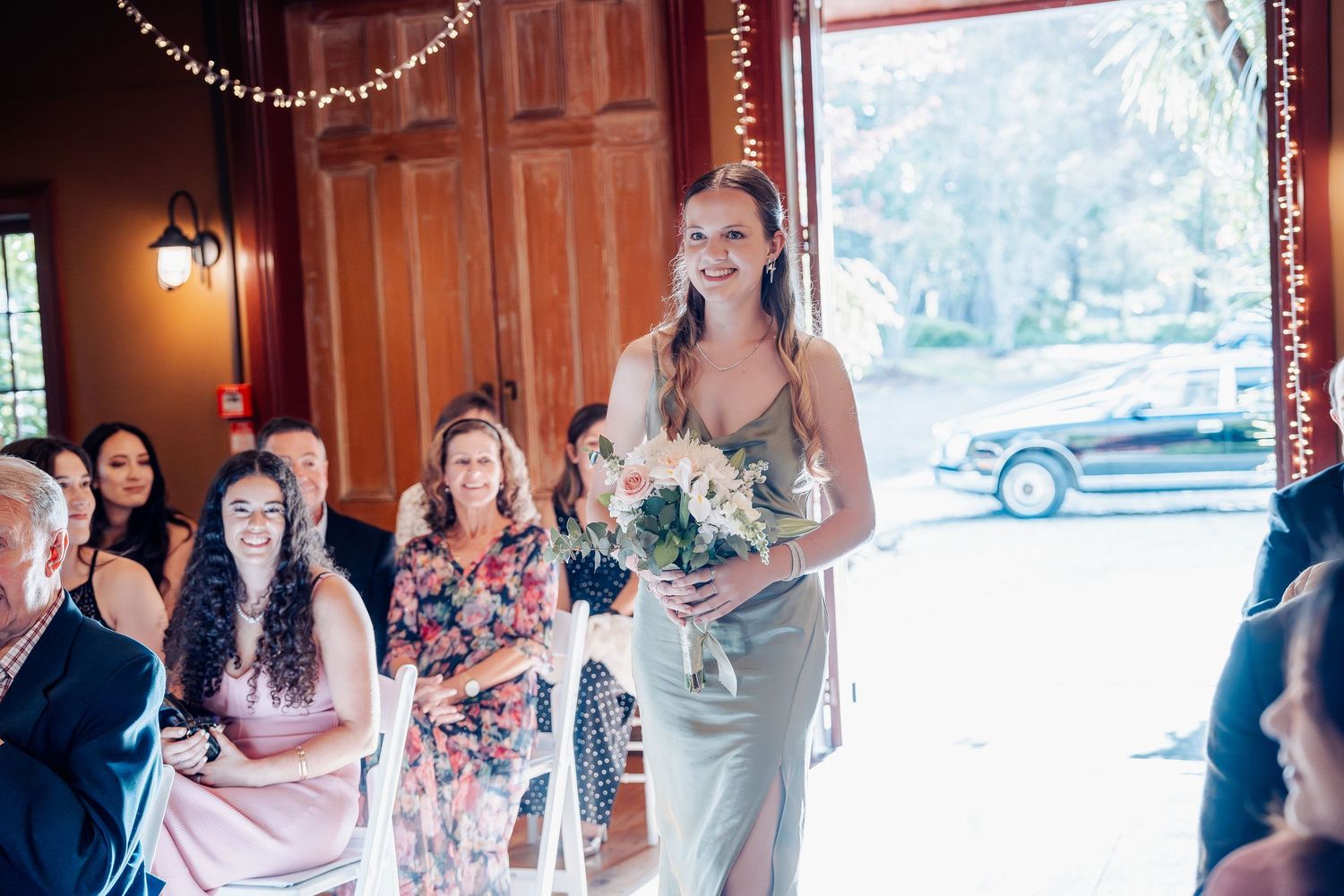 A bride in an elegant white dress carries a teal bouquet during a wedding ceremony at a wood-paneled venue.