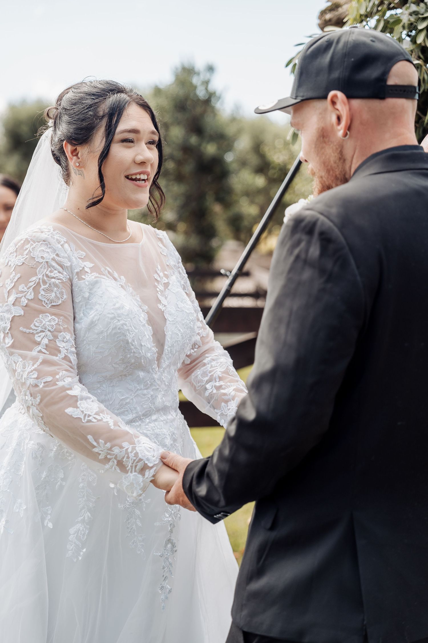 A bride in a lace wedding dress exchanges vows with her partner during an outdoor ceremony.
