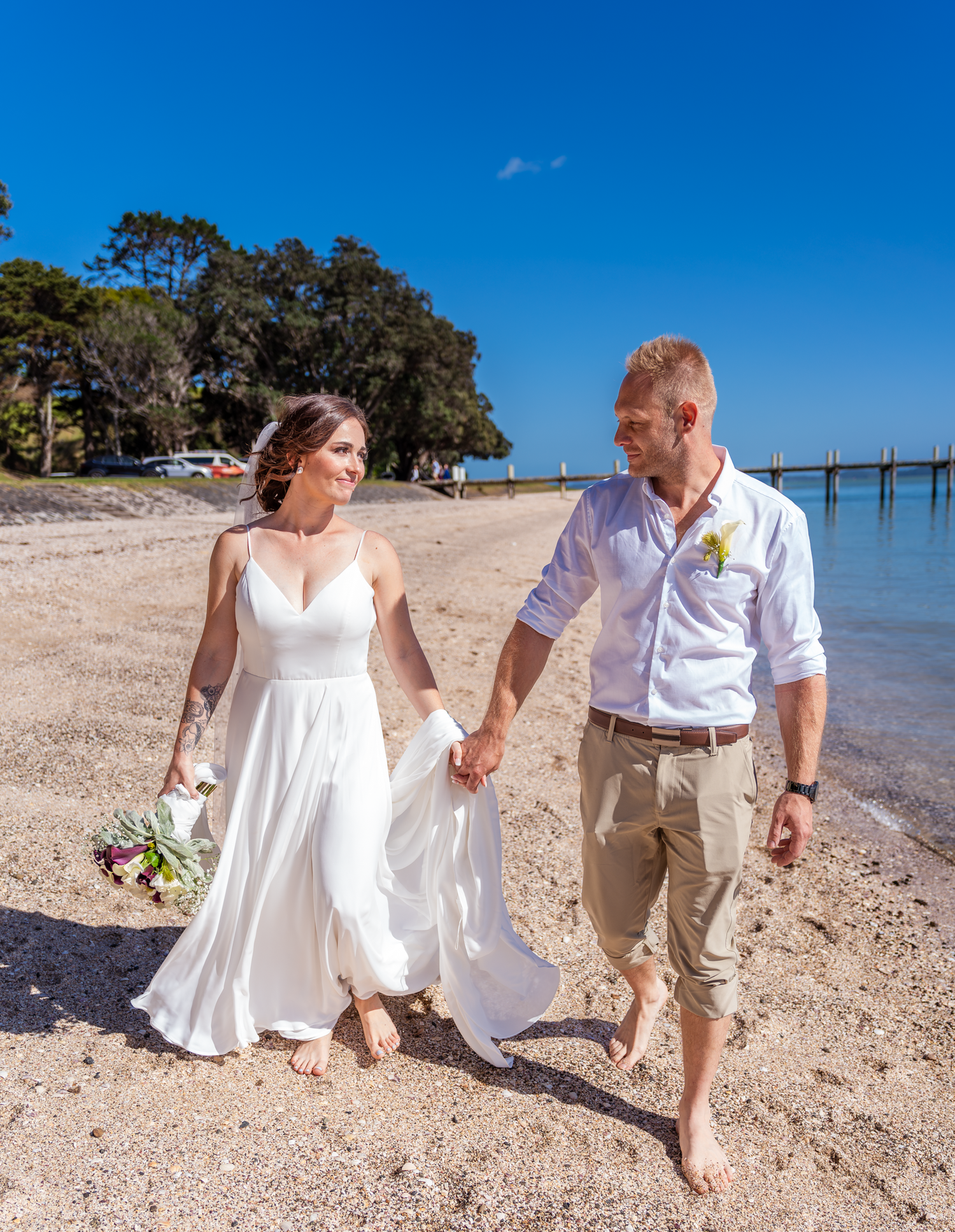 Couple walks barefoot along tropical beach shore holding hands in wedding attire.