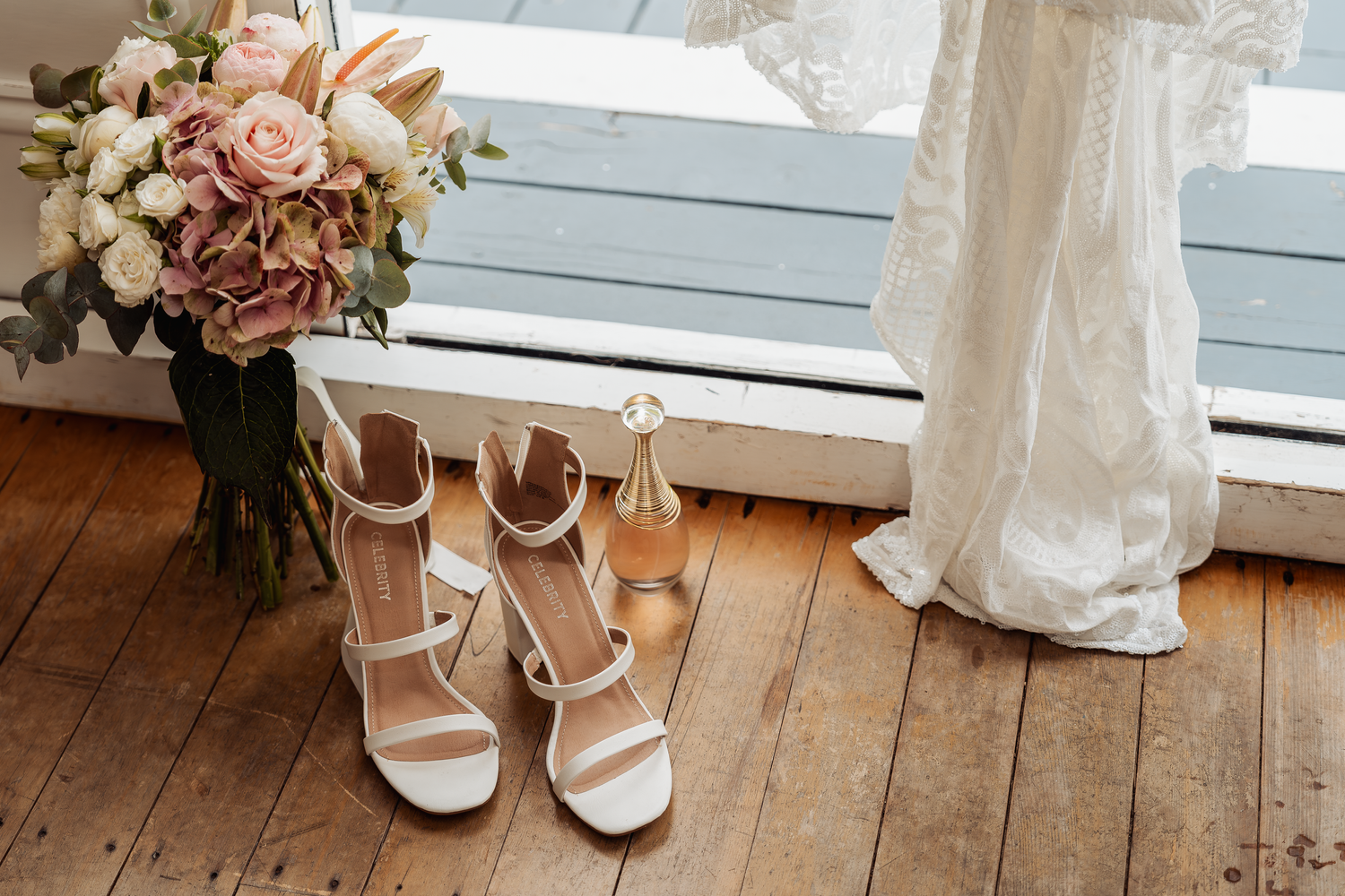 White bridal shoes and pink floral bouquet arranged on wooden floor near window.