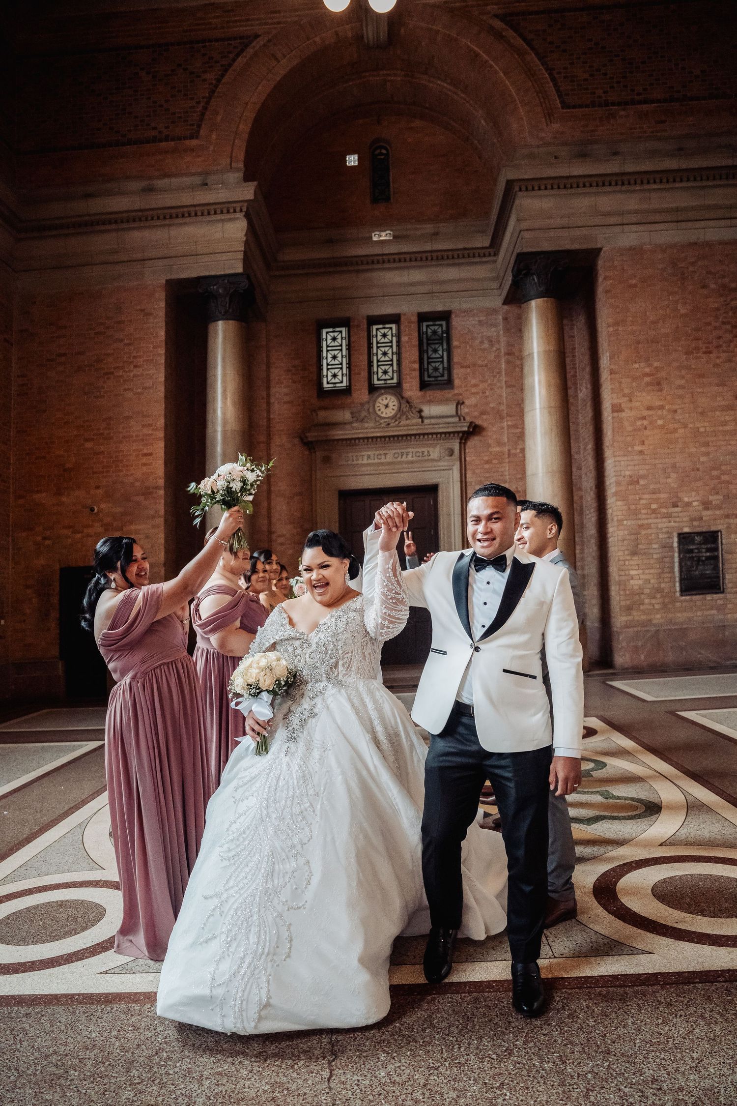 A joyful wedding party smiles and dances together in an elegant brick-walled venue with ornate flooring.