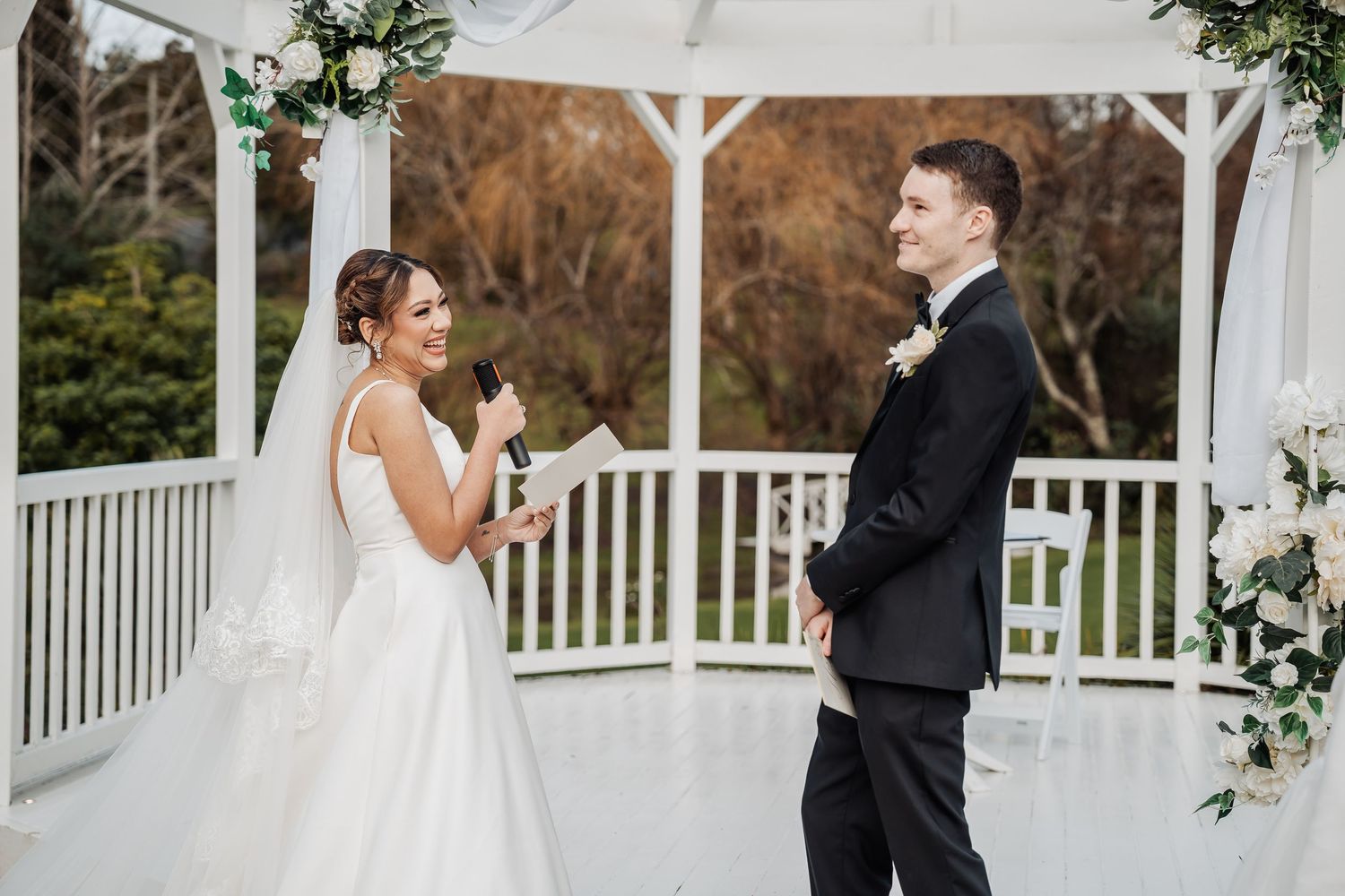 Wedding ceremony taking place under a white wooden gazebo with floral arrangements and winter trees in background.