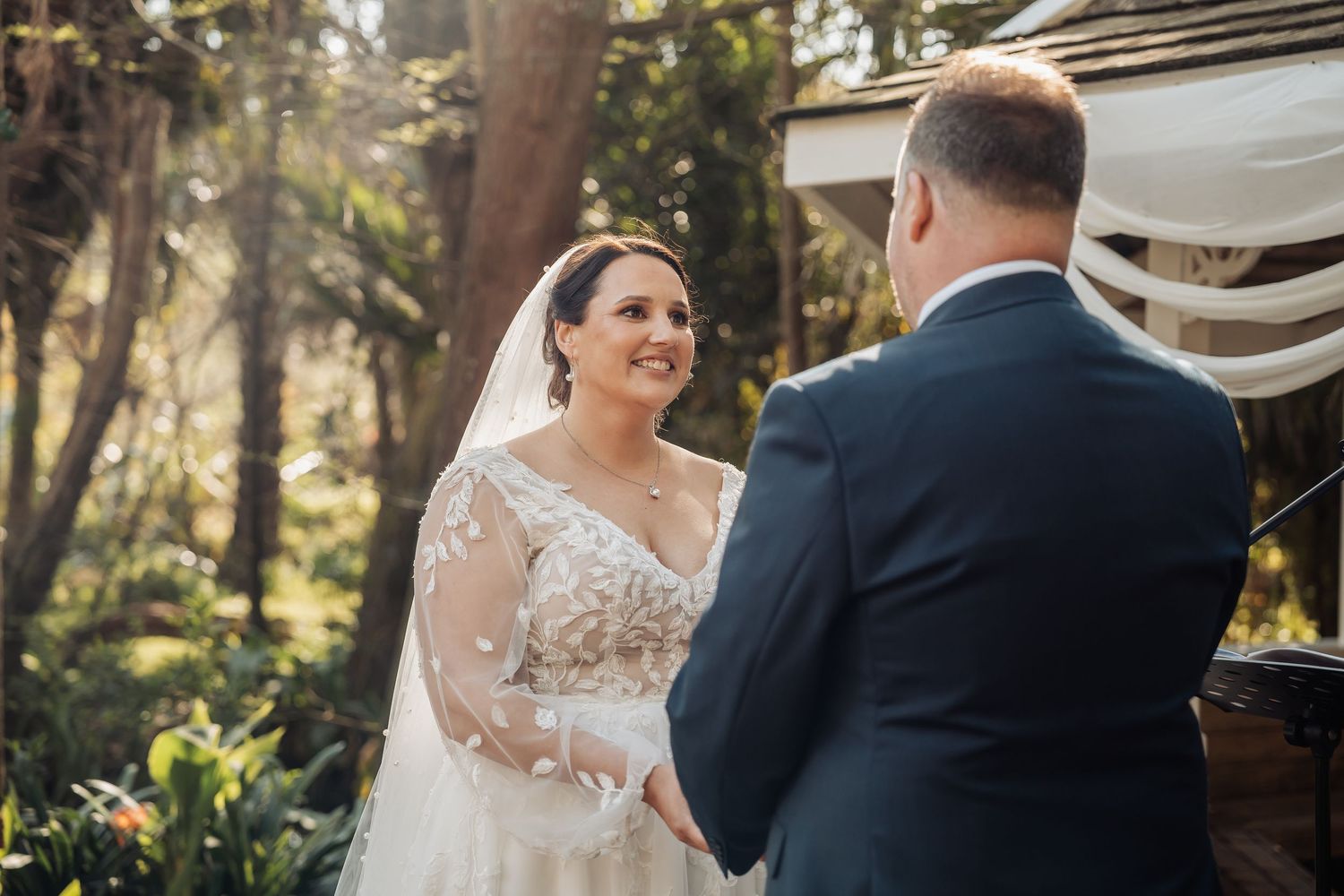 A bride and groom share an intimate moment together during their outdoor wedding ceremony in a sunlit forest setting.