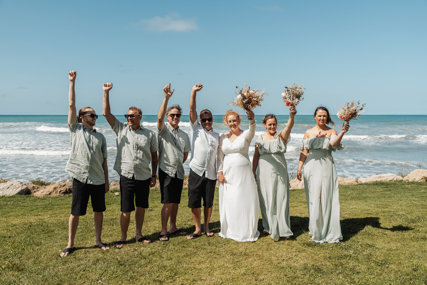 Wedding party celebrates on grassy oceanfront with raised arms and joyful expressions.