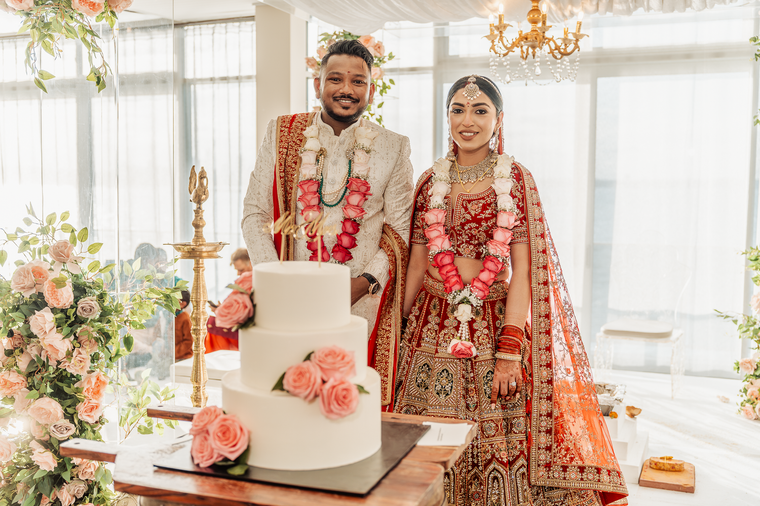 Indian wedding couple in traditional attire posing beside their elegant white tiered cake.