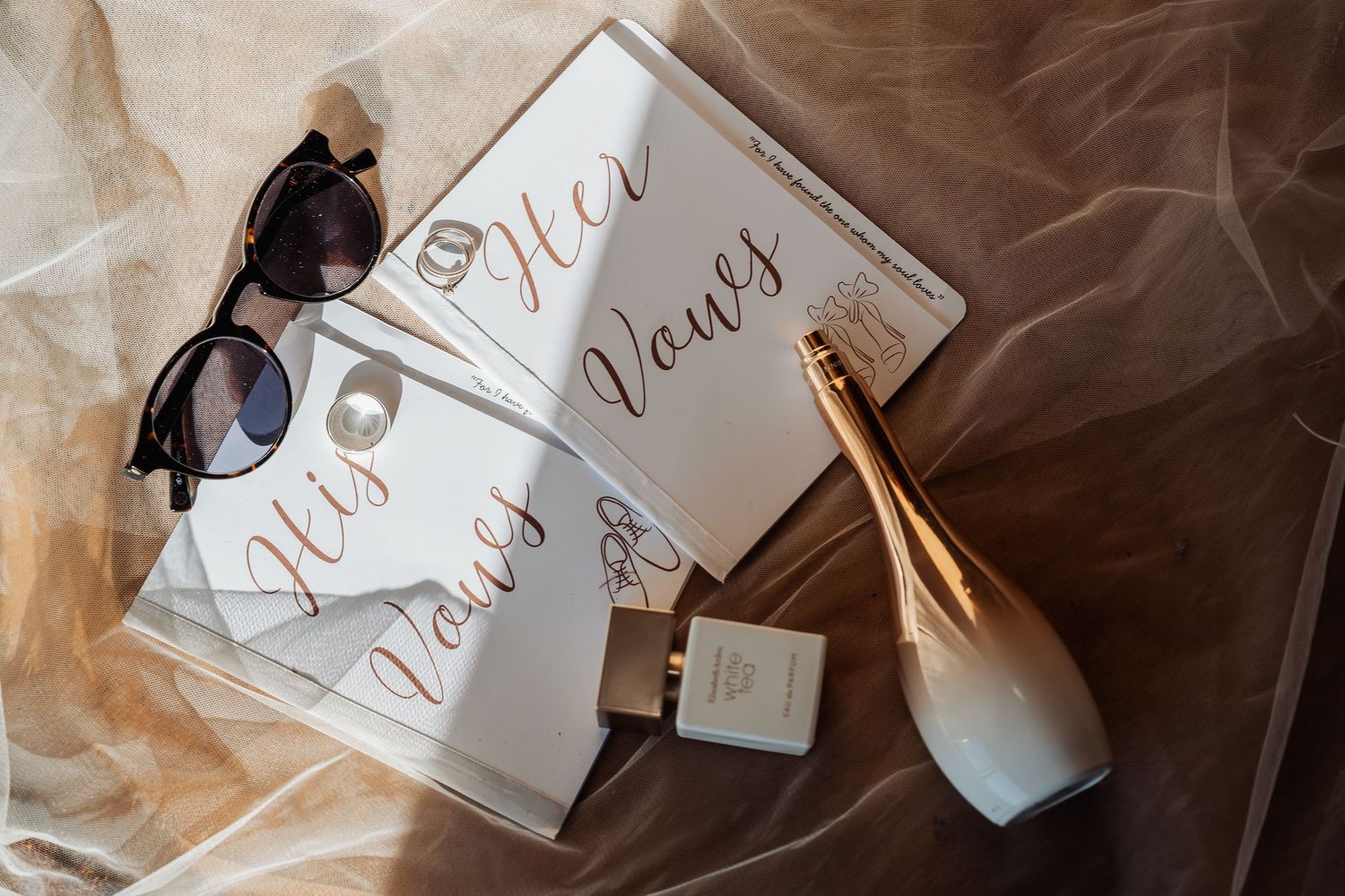 Wedding flat lay with His and Her vows books, sunglasses, perfume, and champagne bottle on tulle.