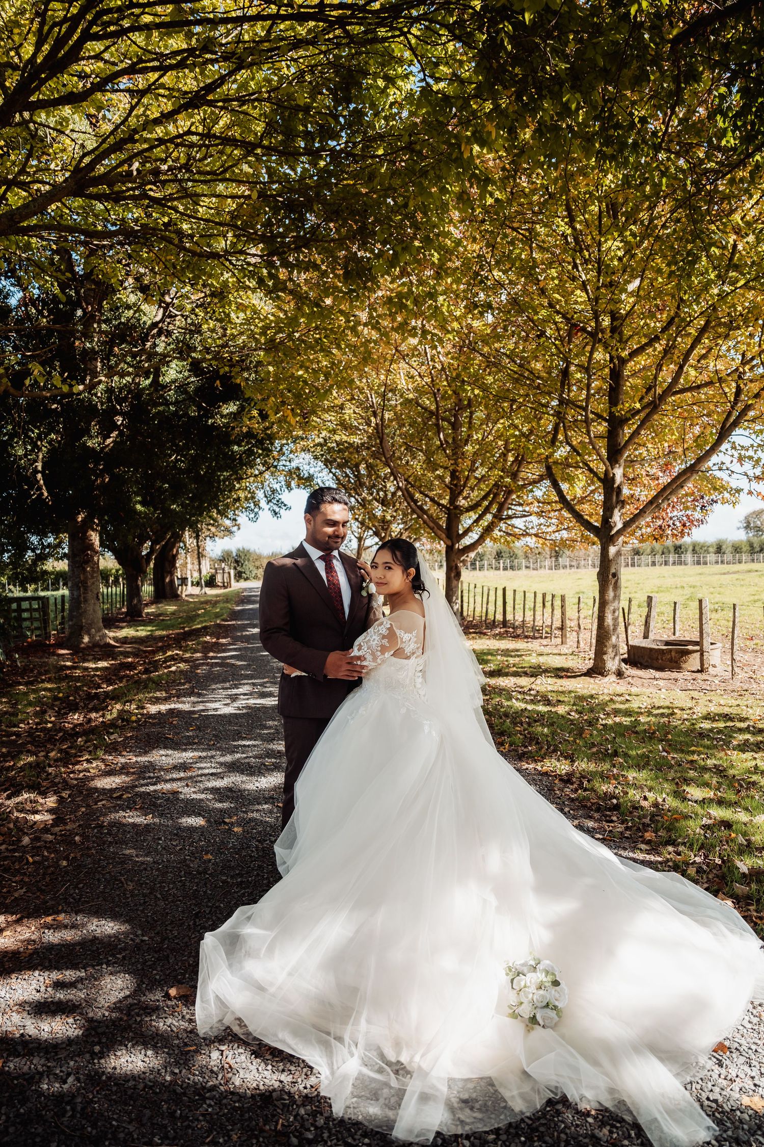 Bride and groom walking hand in hand along a tree-lined autumn path in wedding attire.