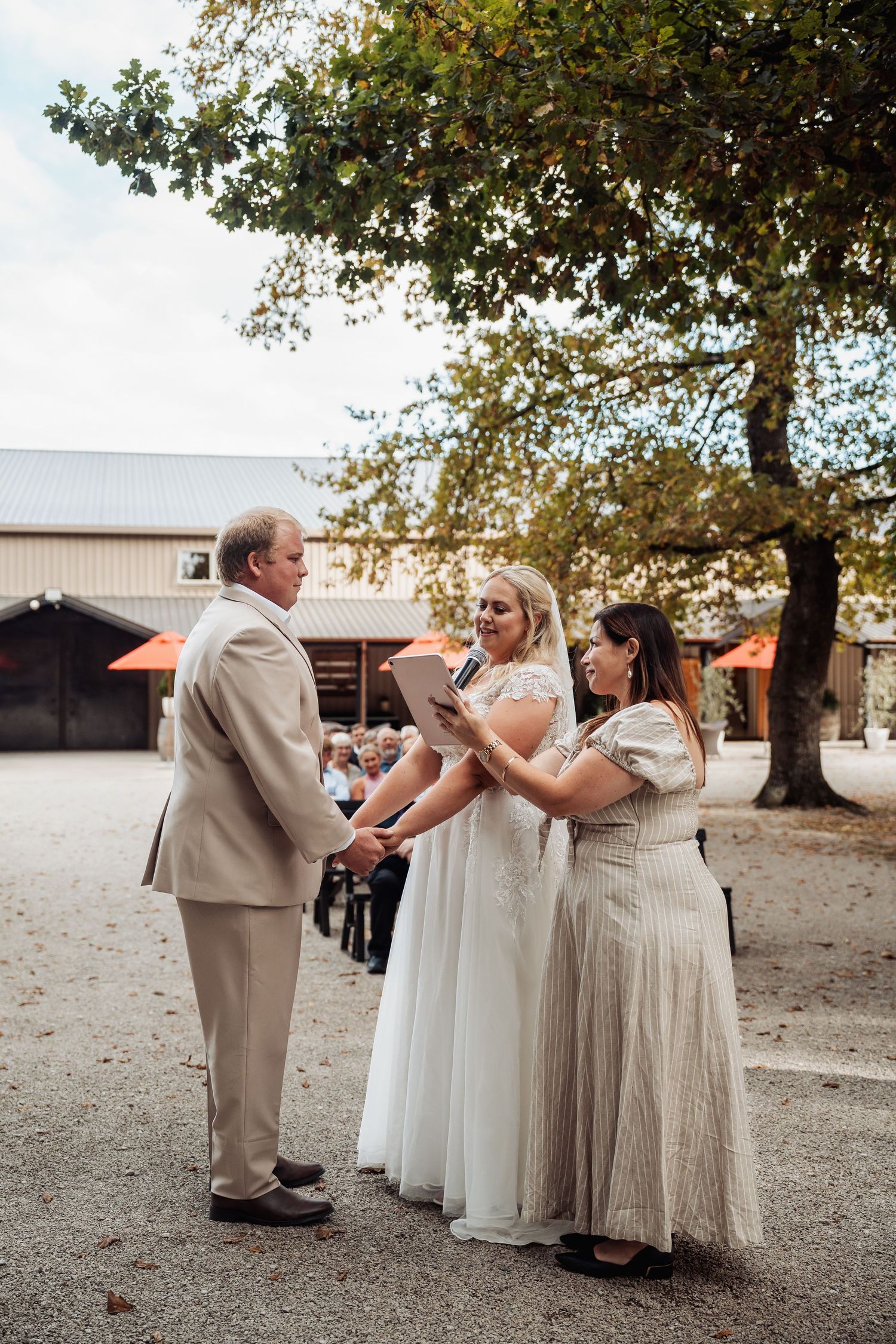 Bride and groom exchange vows outdoors with officiant at rustic barn wedding venue under large tree.