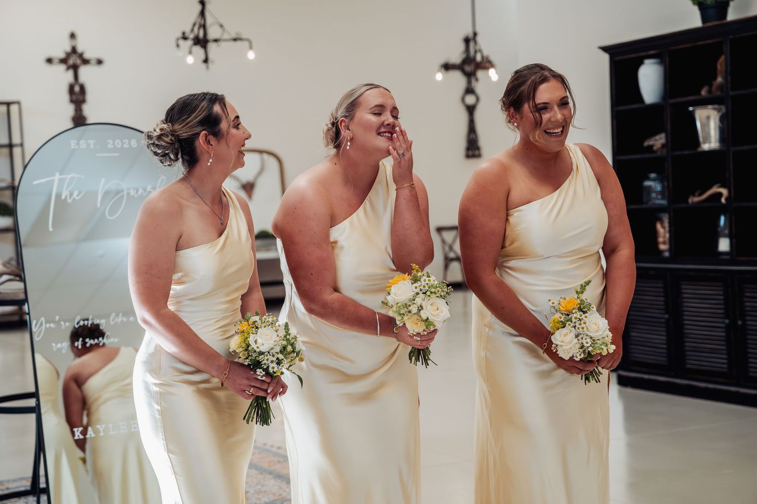 Three bridesmaids in ivory one-shoulder gowns laughing and holding bouquets at wedding ceremony.