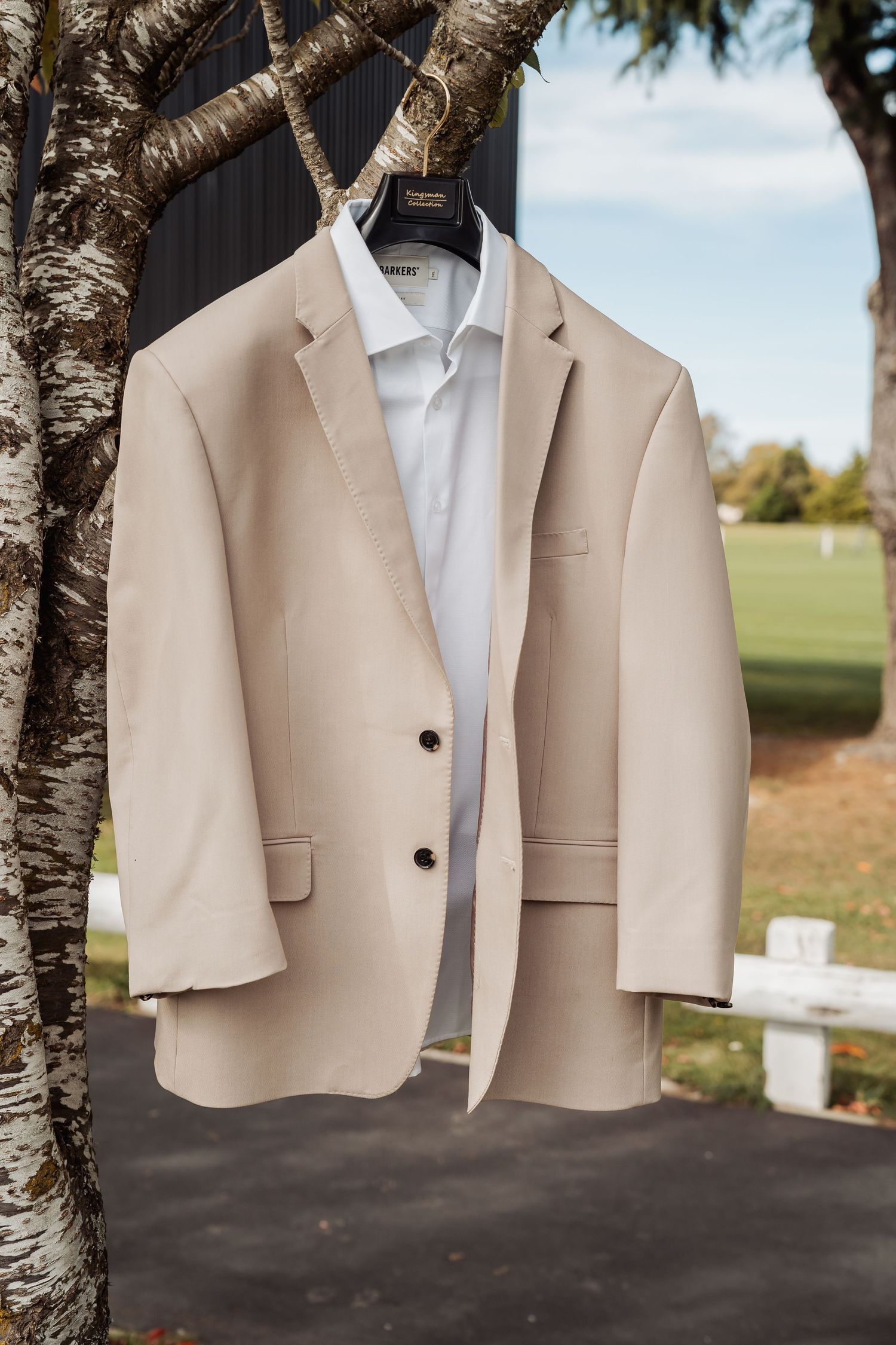 Beige wedding suit jacket hanging on a tree branch outdoors with a white dress shirt.