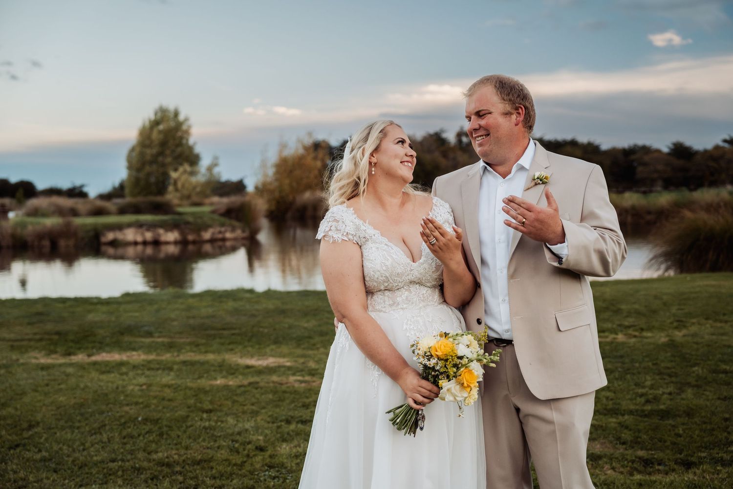 Bride holding yellow bouquet stands with groom in tan suit by a pond at sunset.