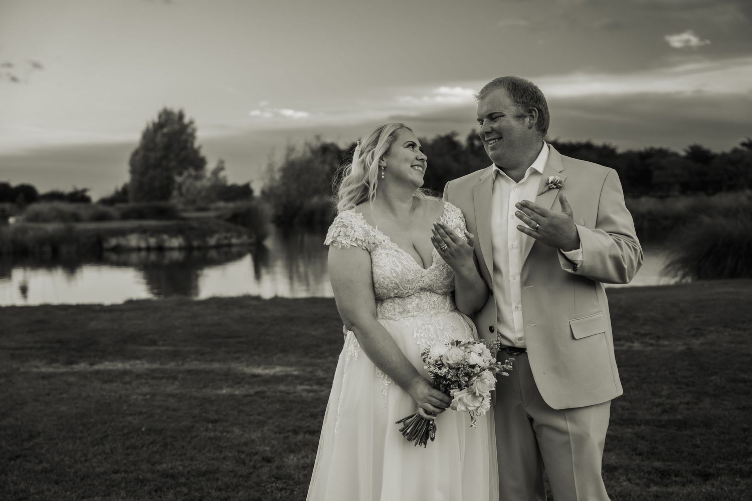 Black and white portrait of bride and groom standing by lakeside at sunset after wedding ceremony.