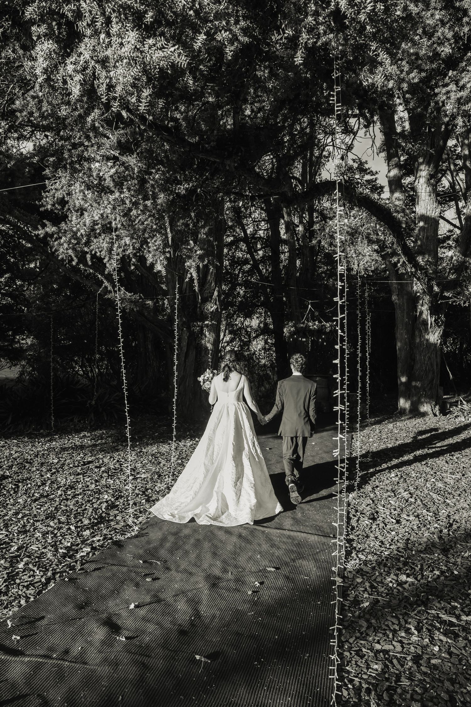 Black and white photo of bride and groom walking hand in hand under fairy lights.