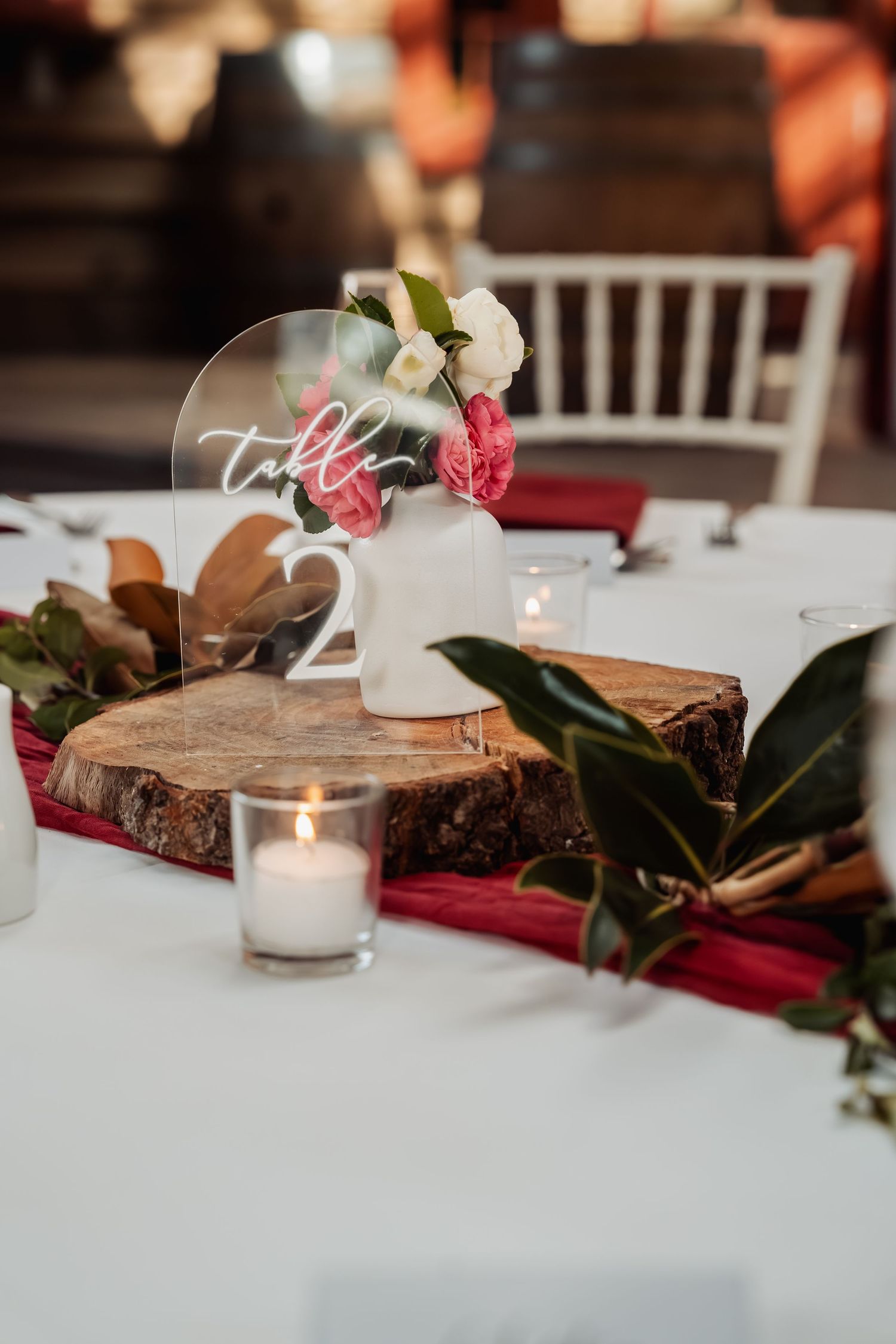 Rustic wedding table centerpiece with wood slice, candles, pink flowers, and acrylic table number sign on white linen.