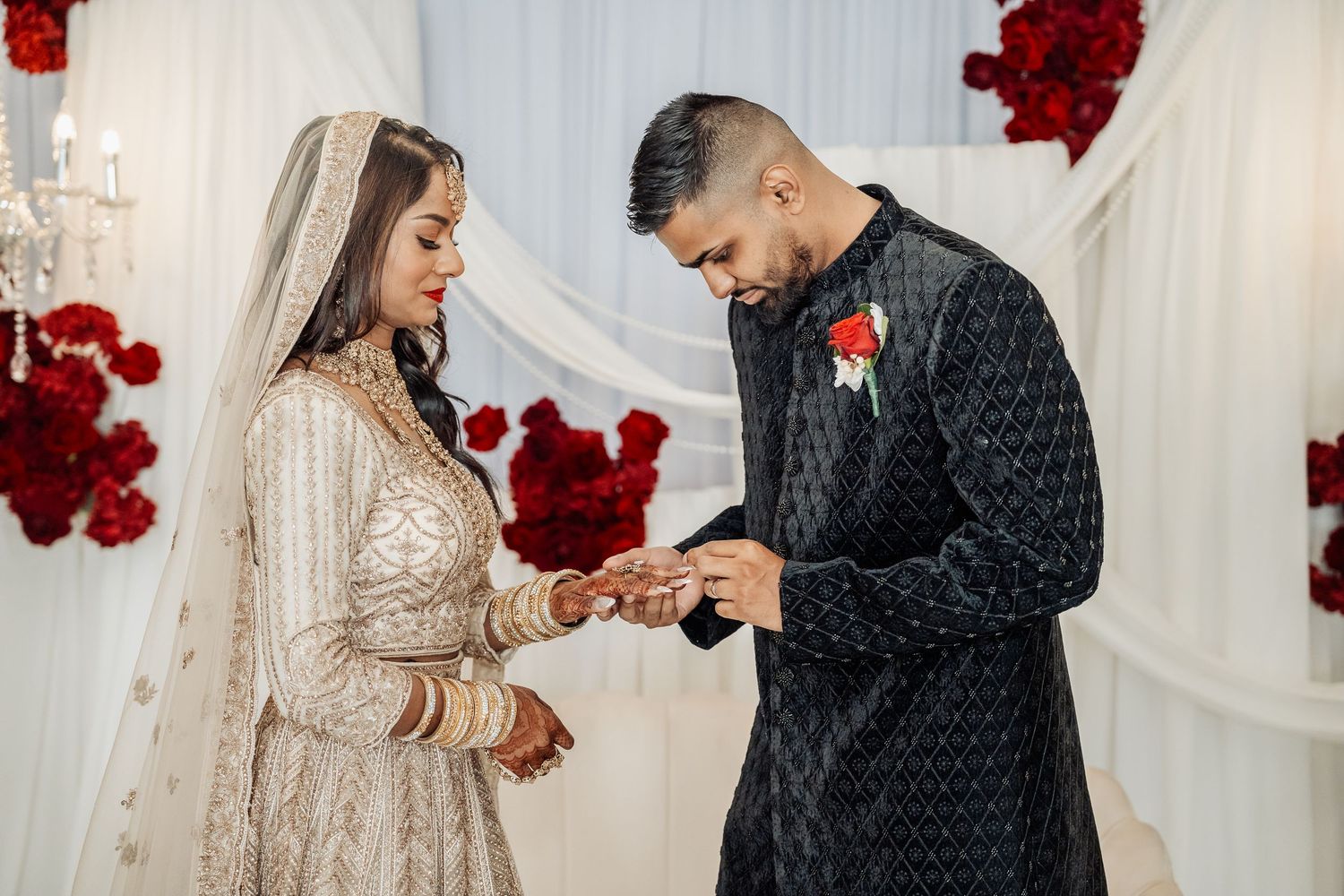A couple exchanges rings during their wedding ceremony against a backdrop of red roses and white drapes.