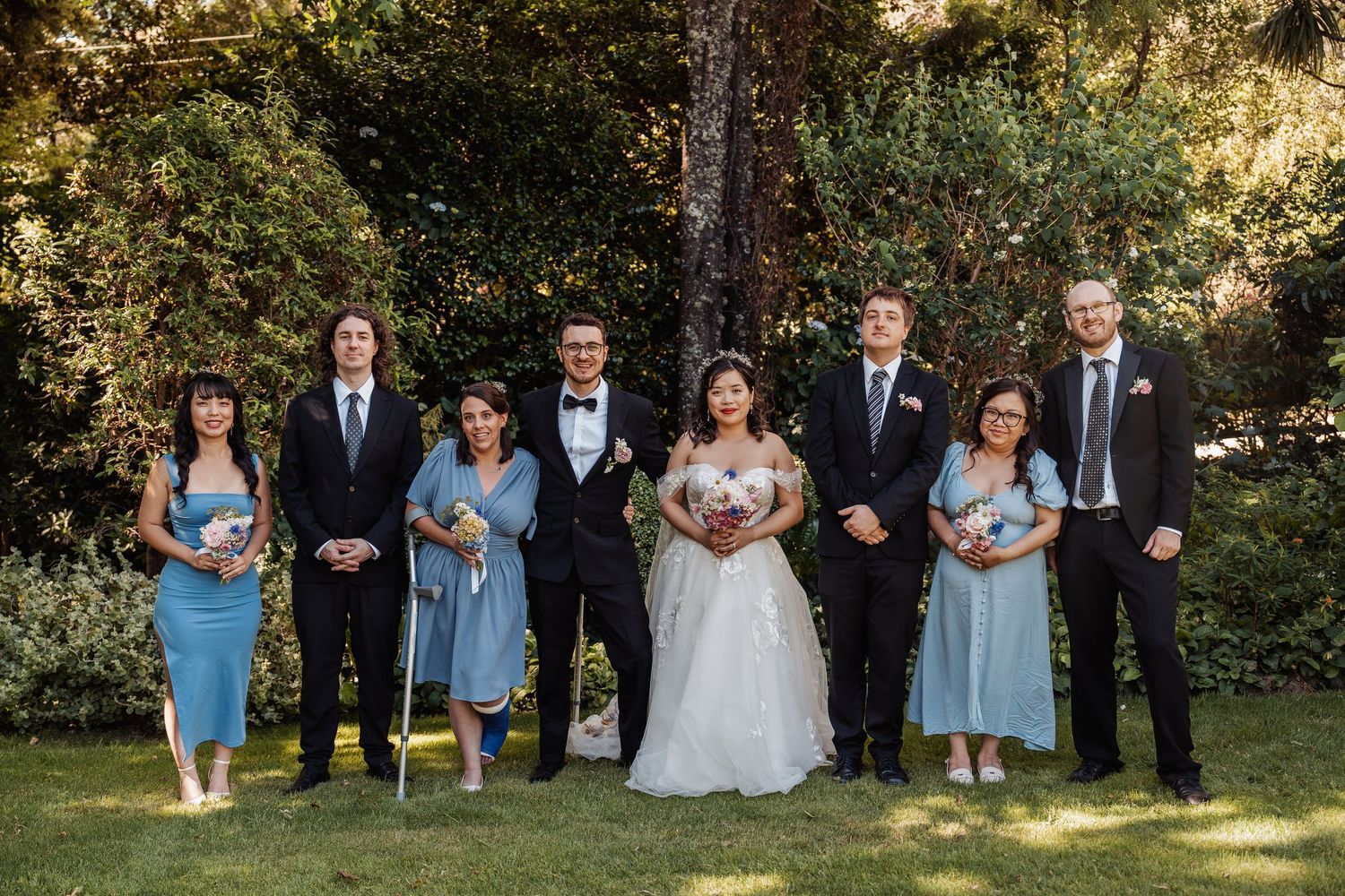 Wedding party poses outdoors in garden, bride in white gown with bridesmaids in blue dresses and groomsmen in black suits.