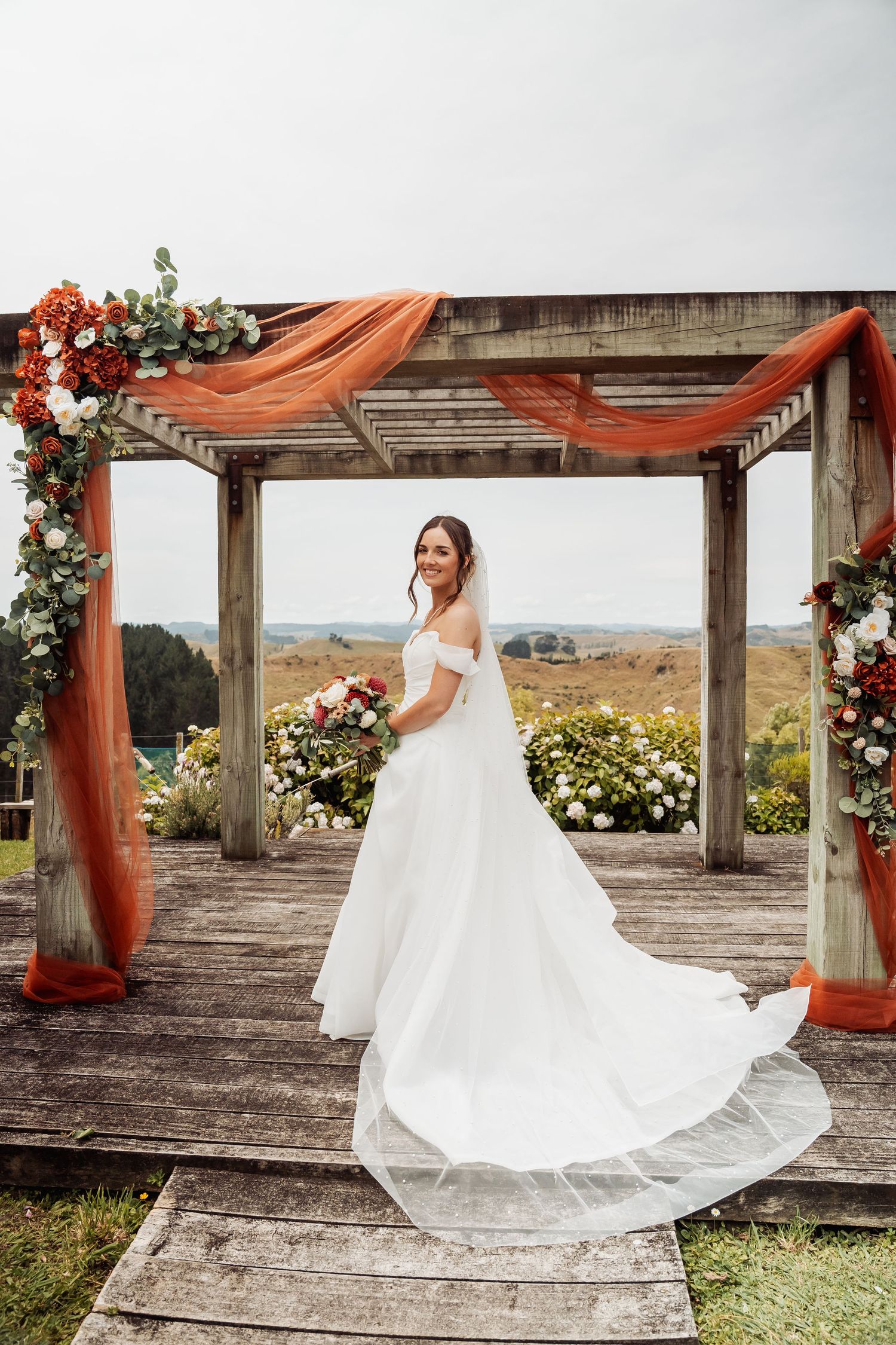 Bride in off-shoulder white gown poses under rustic wooden arch with orange draping and bouquet.