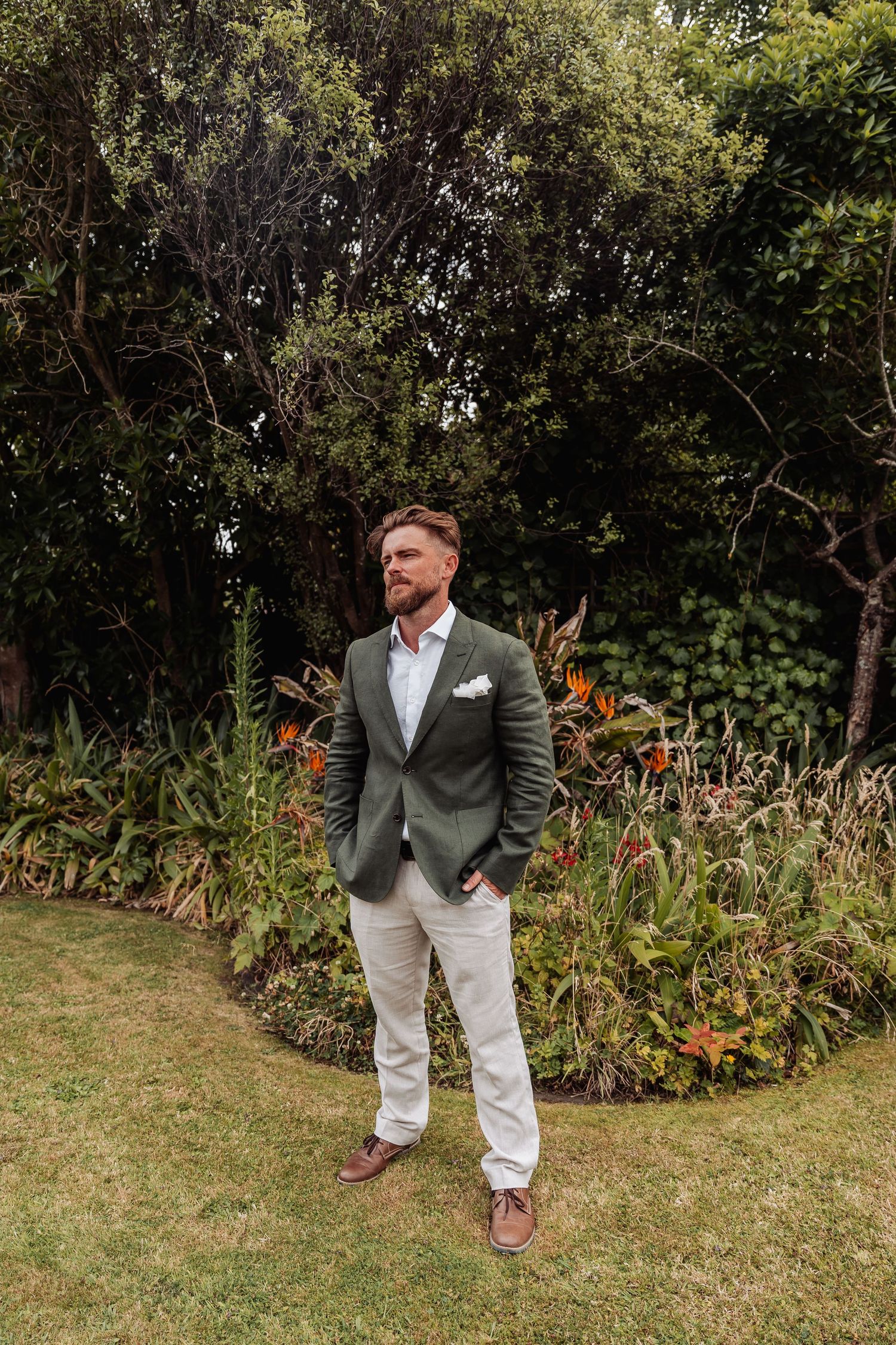 Groom in green blazer and cream trousers stands confidently in a tropical garden.