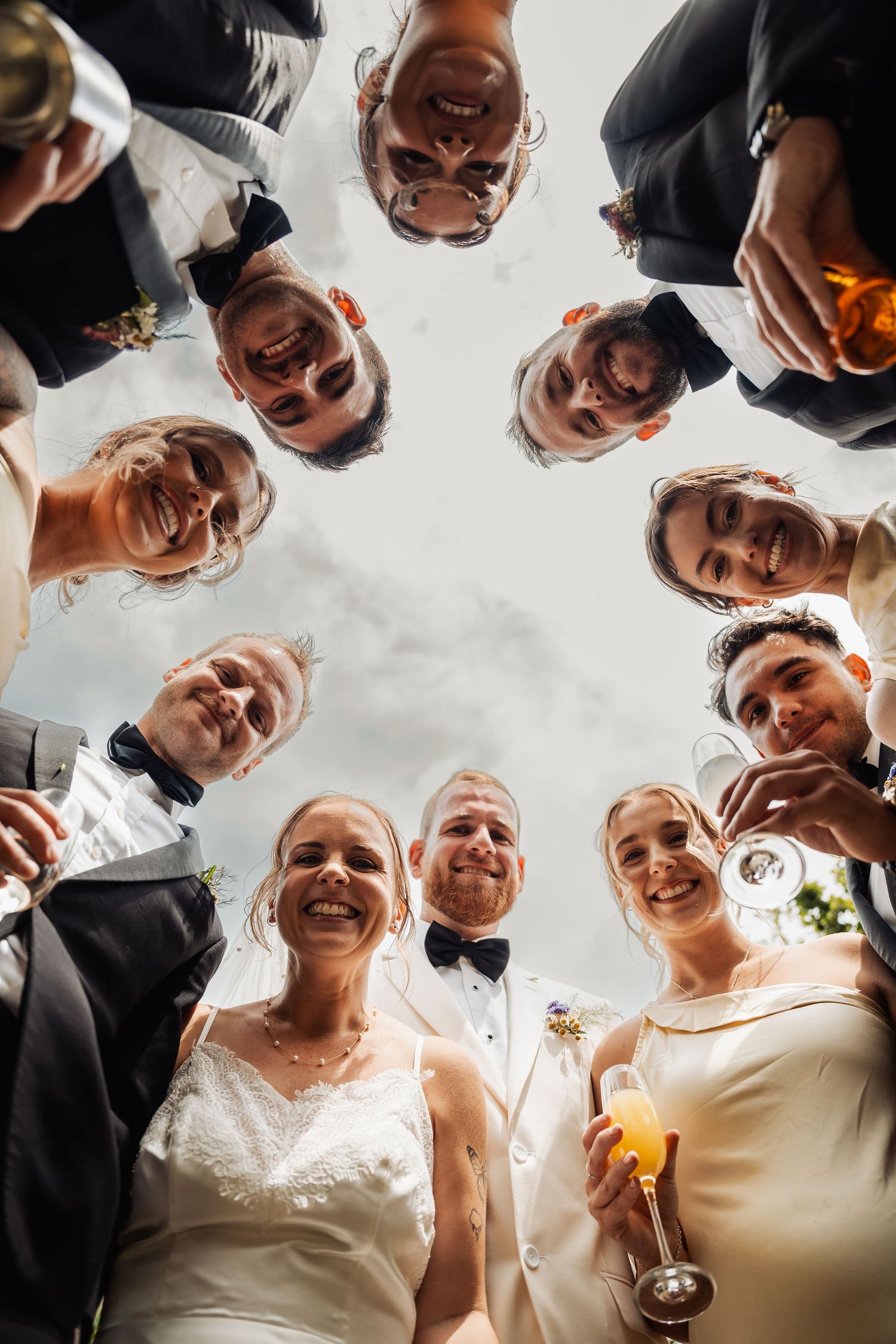 Wedding party celebrating in a circle viewed from below, raising champagne glasses to the sky.