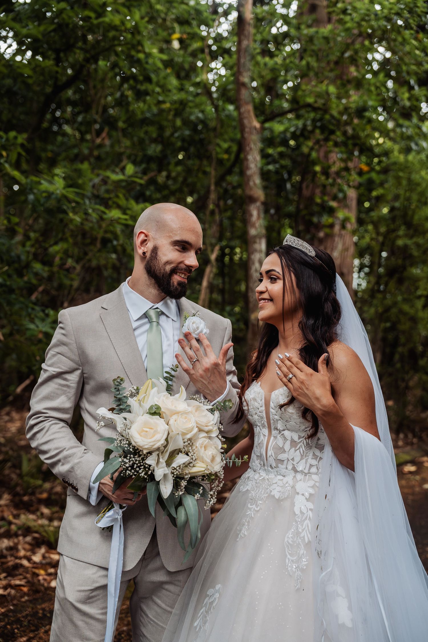 Smiling bride and groom admire their wedding rings in a lush forest setting, groom holding white rose bouquet.