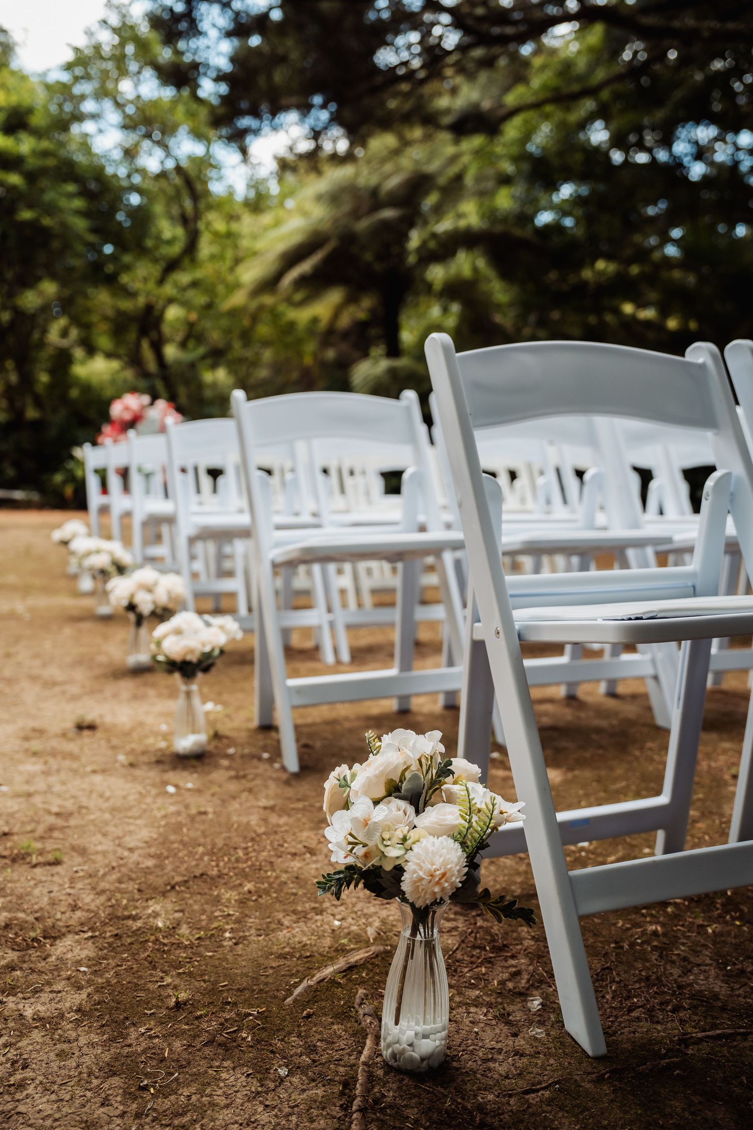 White folding chairs line a garden wedding aisle, adorned with white floral bouquets on a rustic dirt path.