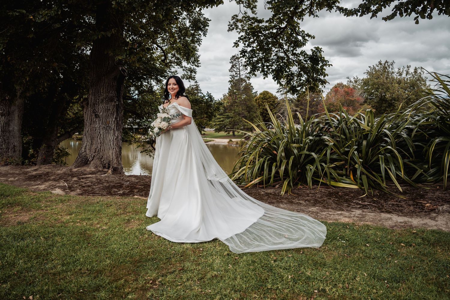 Bride in white gown with long train poses by pond in scenic garden setting.