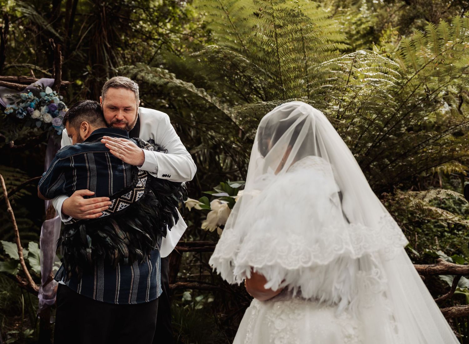 Emotional wedding ceremony moment with groom hugging guest as bride in white gown and veil stands nearby in lush garden.