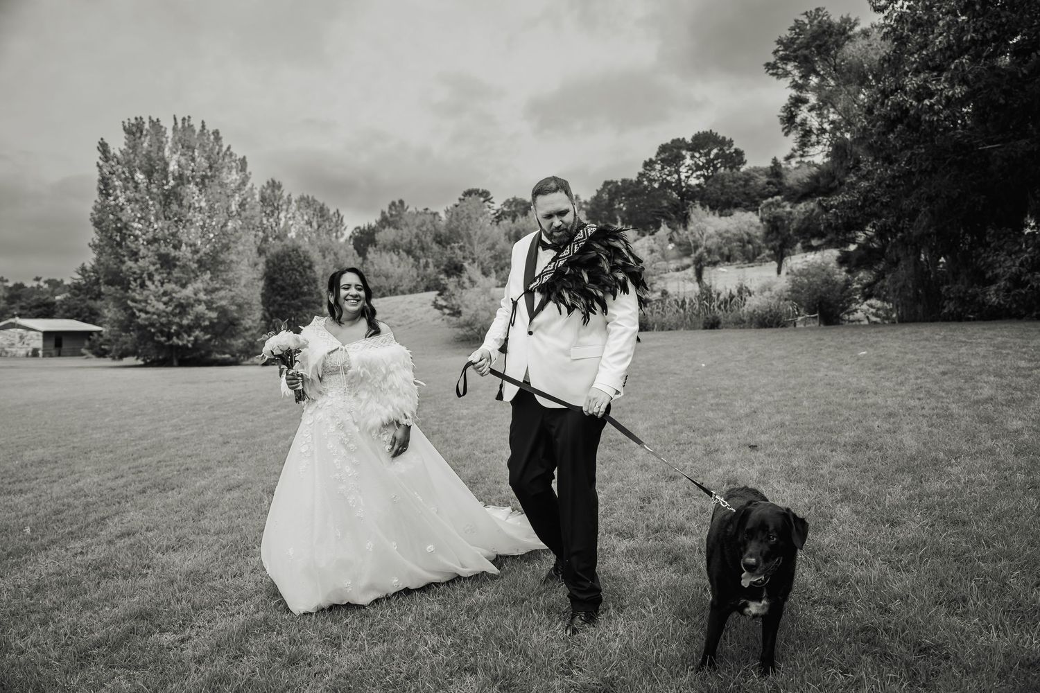 Newlyweds walk their black dog across green field in black and white outdoor wedding portrait.