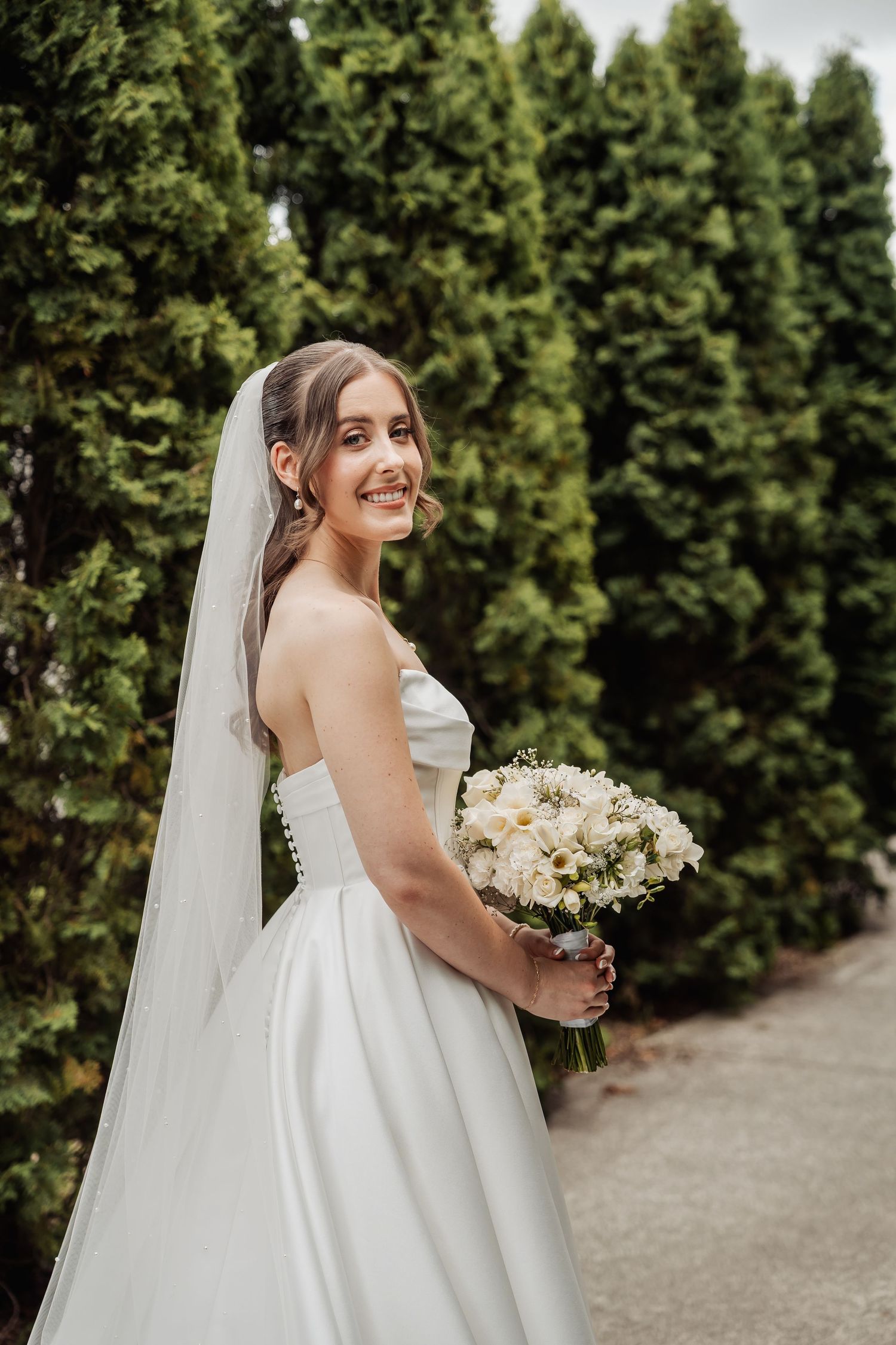 Smiling bride in strapless white gown and veil holds white bouquet outdoors with tall green trees behind her.