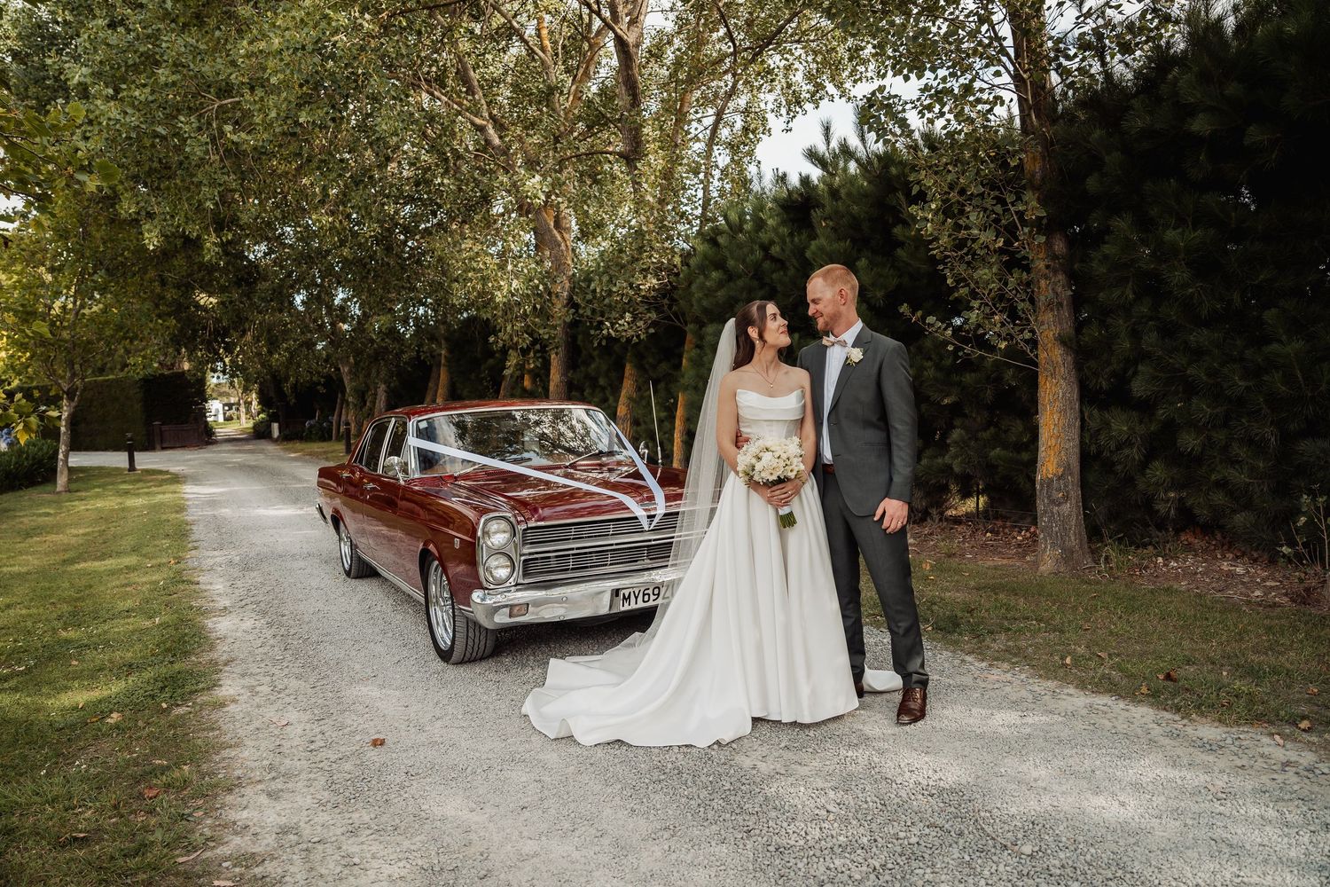 Bride and groom posing beside a classic vintage red car on a tree-lined gravel driveway at their wedding.