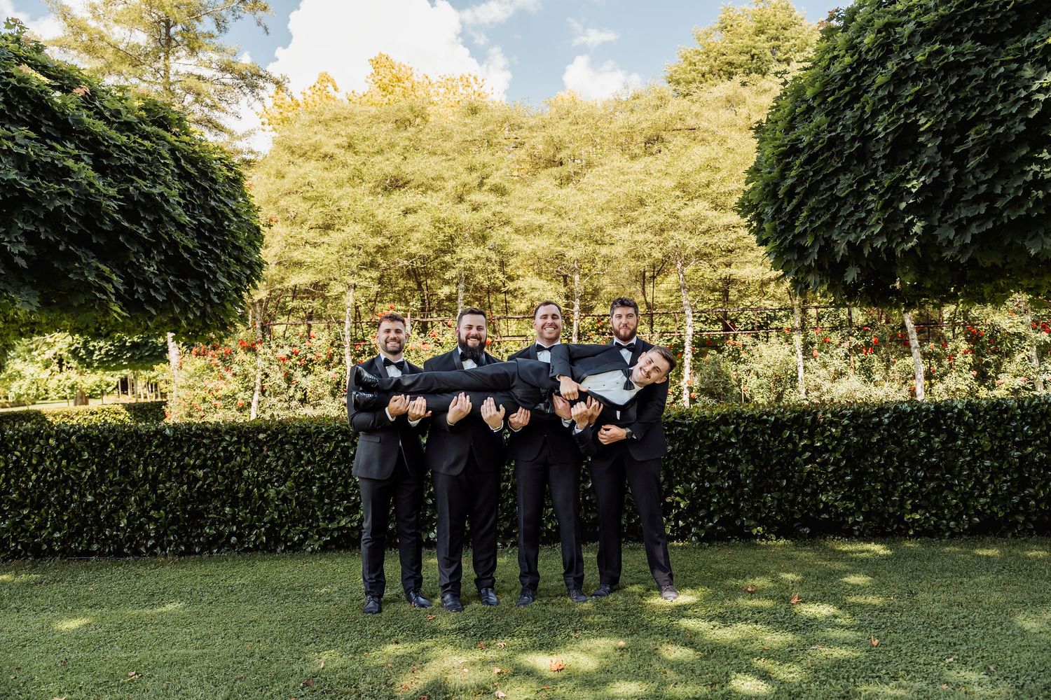 Five groomsmen in black suits laughing and posing together in a lush garden setting.