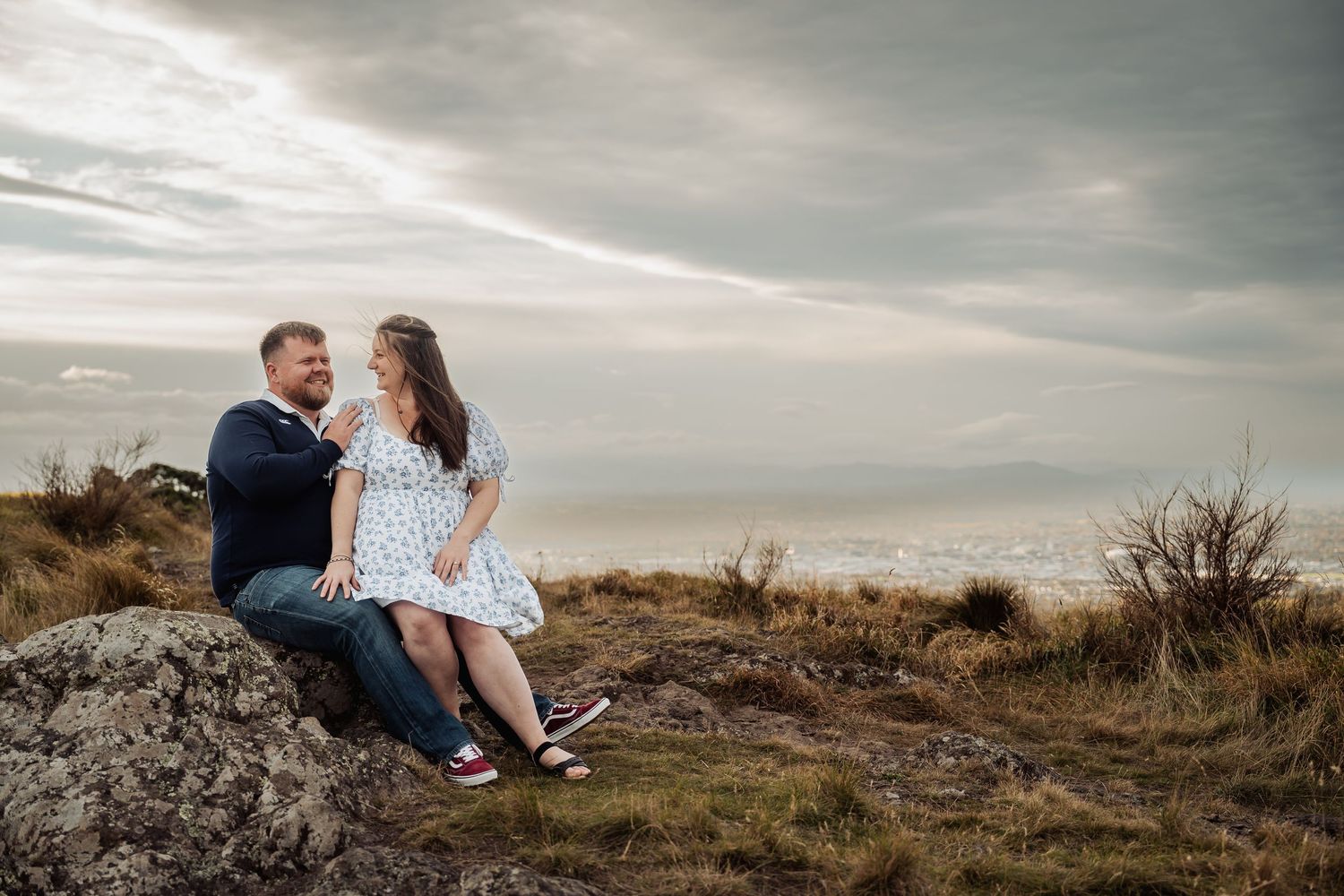 Couple sitting on a rock by the coast, sharing a romantic moment with a scenic ocean and cloudy sky backdrop.